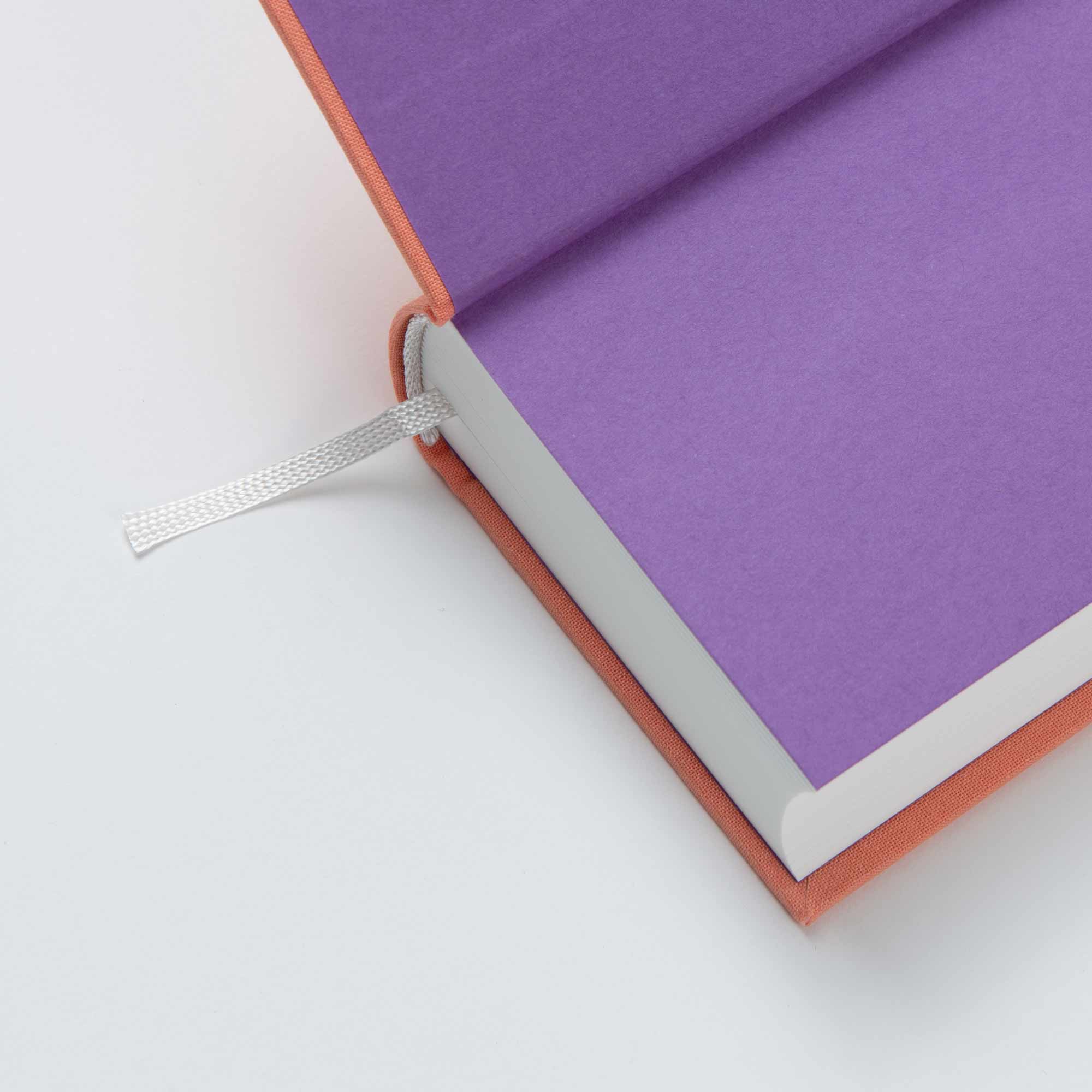 Das OWAD Shine Bright-Notizbuch von paperlux.store mit violetten Seiten, orangefarbenem Einband und weißem Lesezeichenband ruht elegant auf einer makellos weißen Oberfläche.