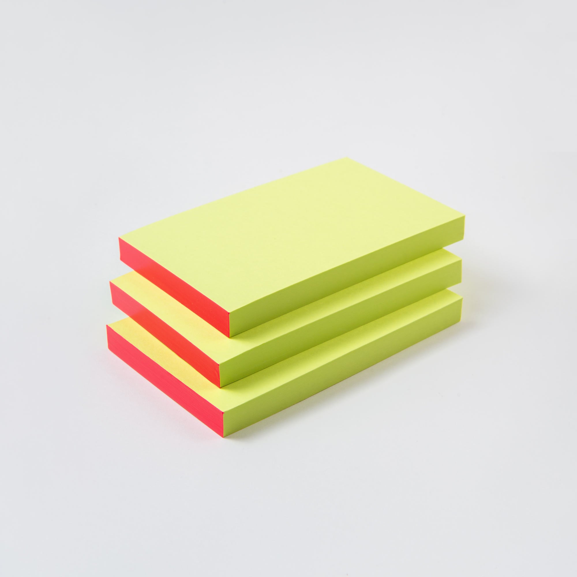 Ein Stapel von vier paperlux Abreissblöcken Neon aus Feinstpapier-Resten, mit gelben Spitzen und fluoreszierenden rosa Rändern, ordentlich auf einem einfarbig weissen Hintergrund angeordnet.