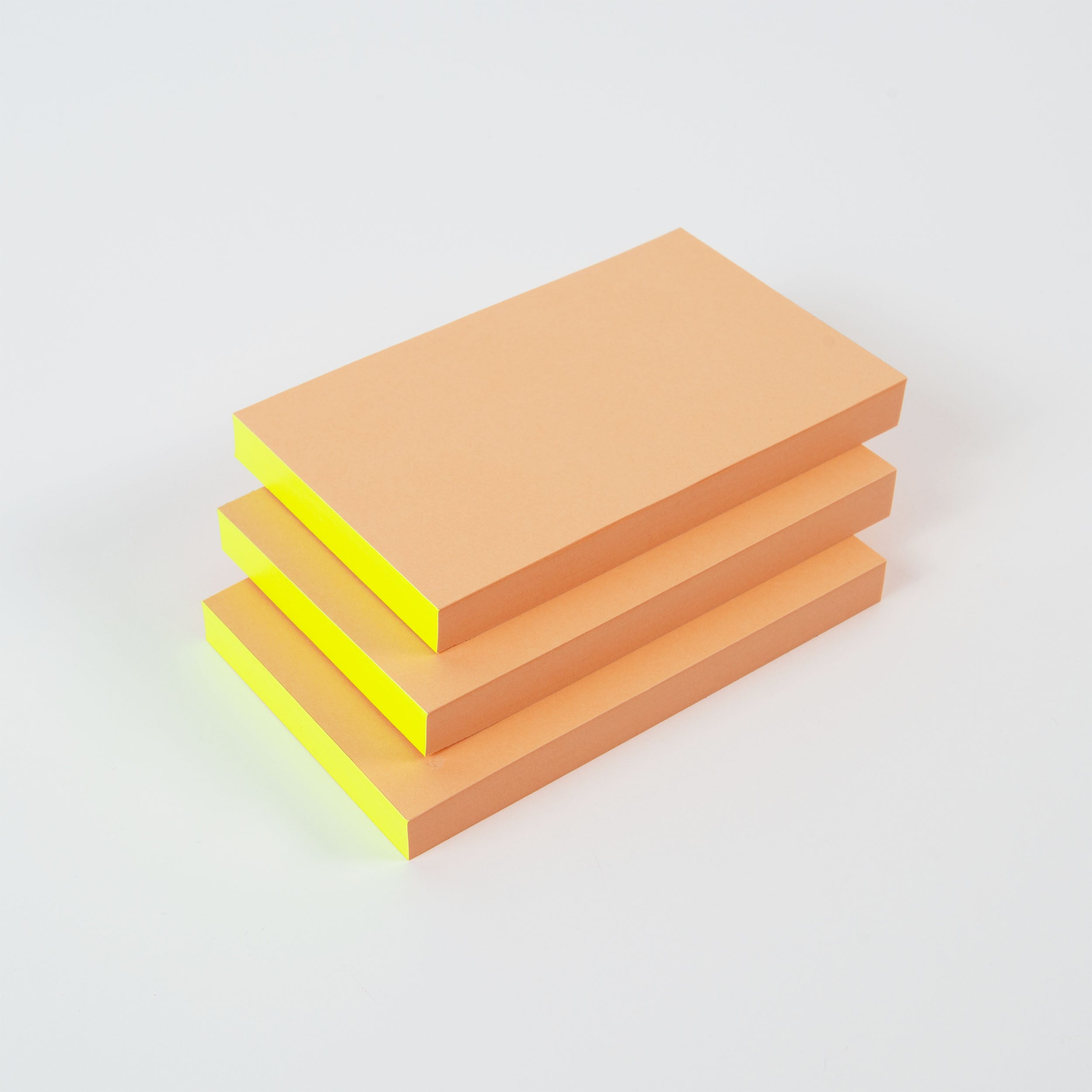 Drei Stapel paperlux Abreissblöcke Neon, hergestellt aus Feinstpapier-Resten, sind fein säuberlich angeordnet. Die orangefarbenen Zettel mit gelbem Rand heben sich lebhaft von dem schlichten weißen Hintergrund ab.