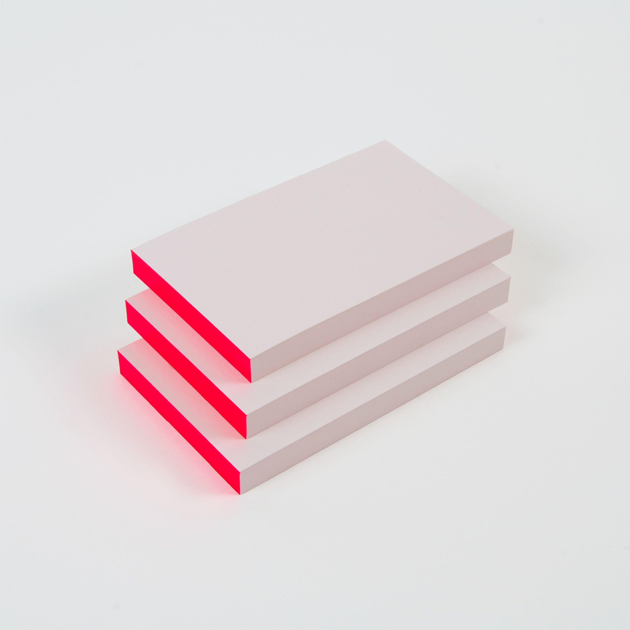 Die Abreissblöcke Neon von paperlux.store bestehen aus drei leicht außermittig angeordneten, weißen rechteckigen Blöcken mit rötlich gefärbten Kanten. Sie liegen auf einem sauberen, weißen Hintergrund, der an die reine Qualität von Feinstpapier-Resten erinnert.
