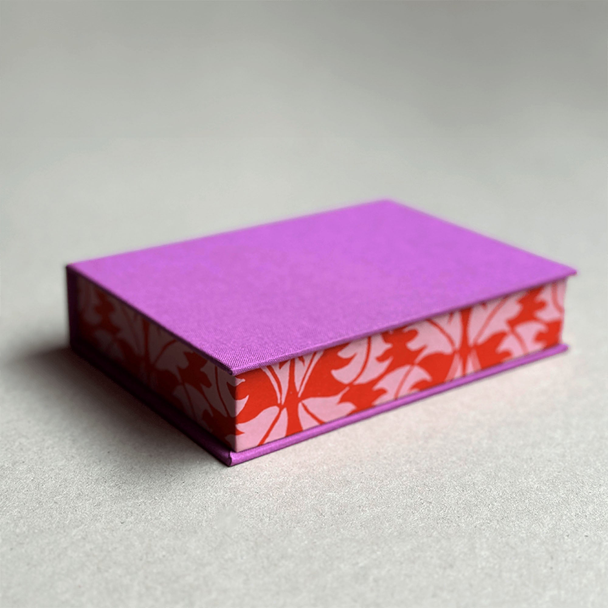 Der Workshop Clamshell Box von paperlux.store ist eine geschlossene, rechteckige Box mit violettem Deckel und rot-weißen, floral gemusterten Seiten, die auf einer neutralen grauen Oberfläche gezeigt wird.