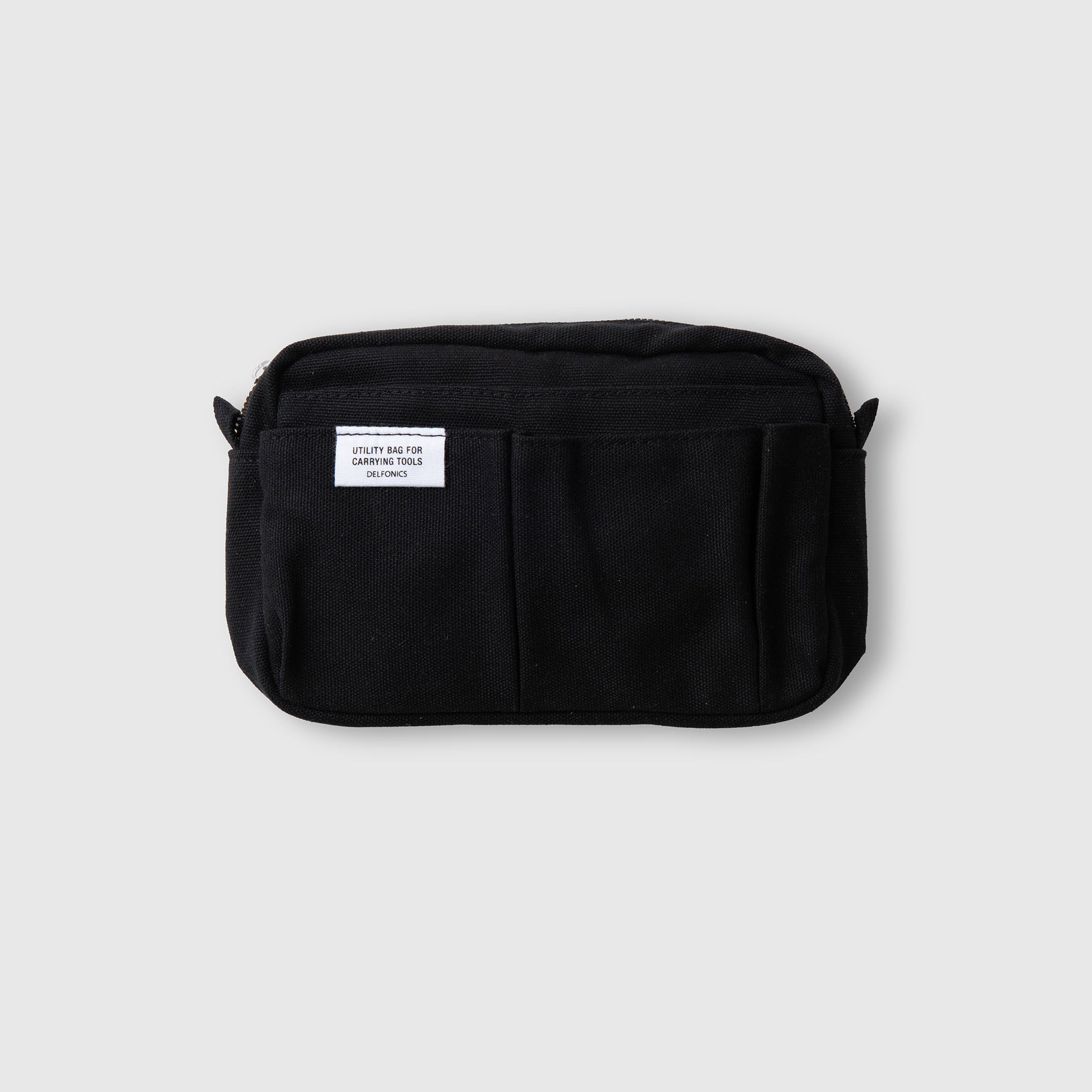 Ein kleiner schwarzer Stoffbeutel, der „Delfonics Inner Carrying Bag S“, mit zwei Fronttaschen, einem Reißverschluss an der Oberseite und einem weißen Etikett mit der Aufschrift „UTILITY BAG 019 LINEN TOOLS CONTAINER“. Der Beutel wird vor einem schlichten hellgrauen Hintergrund präsentiert.