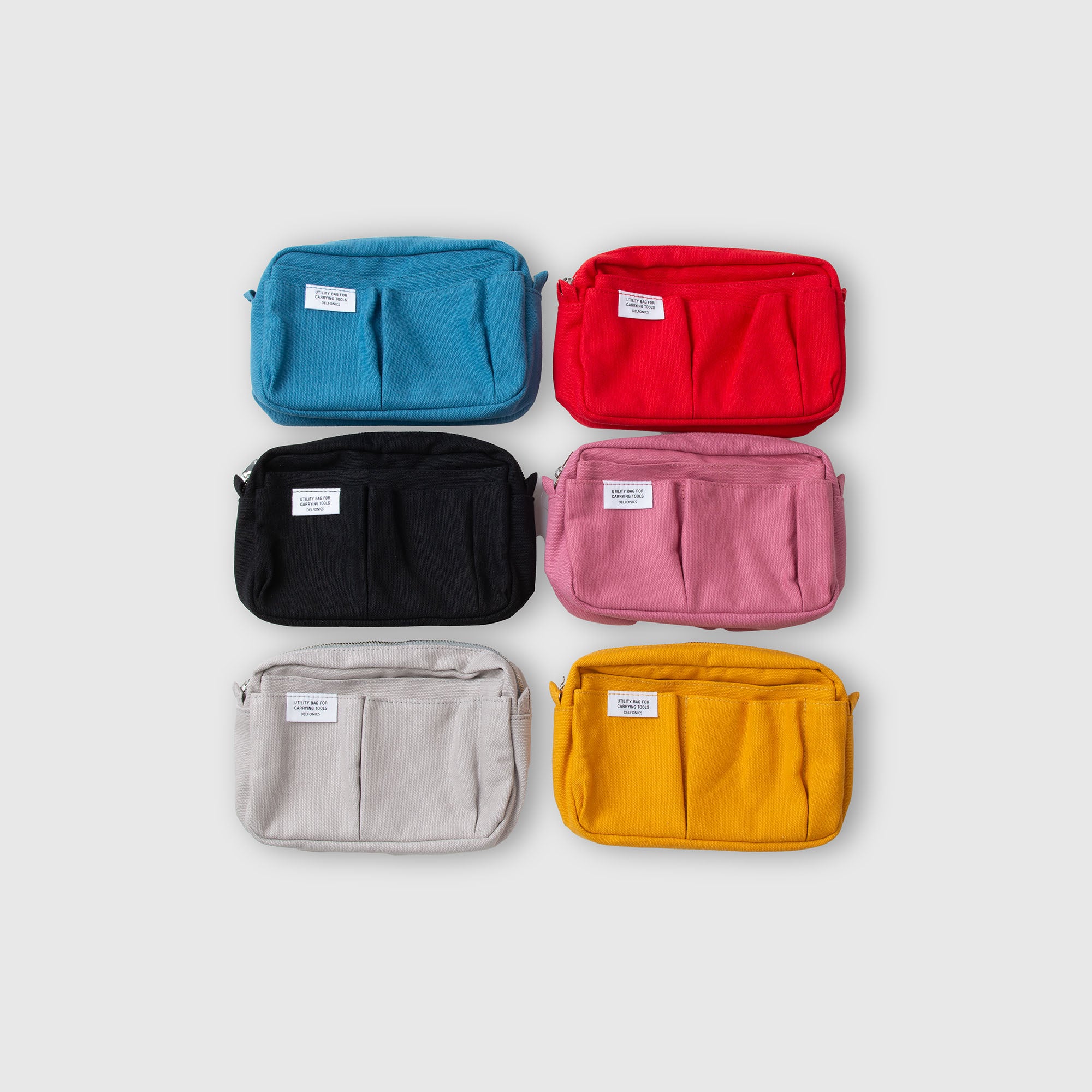 Sechs „Inner Carrying Bag S“-Taschen von Delfonics – ideal als Organizer-Tasche – sind in zwei Reihen zu je drei Stück vor einem schlichten hellgrauen Hintergrund abgebildet. Die Baumwolltaschen von Delfonics sind in den Farben Blau, Rot, Schwarz, Rosa, Grau und Gelb erhältlich und verfügen jeweils über zwei Vordertaschen sowie ein praktisches weißes Etikett.