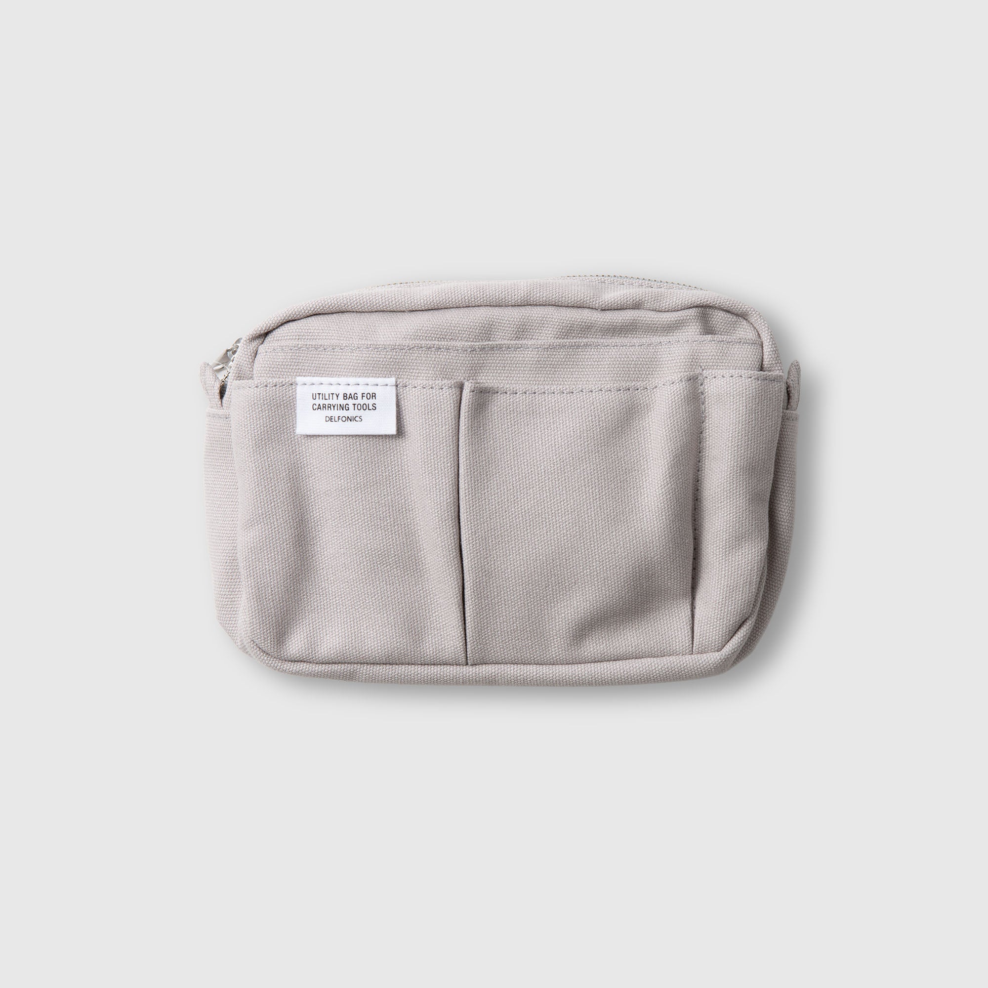 Eine hellgraue Delfonics Inner Carrying Bag S, die den minimalistischen Stil der Marke widerspiegelt, verfügt über zwei Fronttaschen, einen sicheren Reißverschluss und ein kleines weißes Delfonics-Etikett. Diese praktische Organizer-Tasche wird vor einem schlichten, hellen Hintergrund gezeigt.