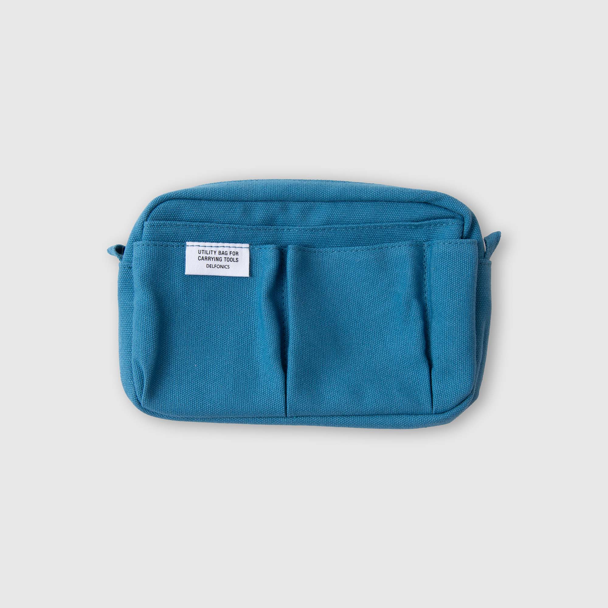 Die „Delfonics Inner Carrying Bag S“ von Delfonics ist eine kleine, rechteckige, blaue Stofftasche mit zwei Vordertaschen und einem weißen Etikett oben links, abgebildet vor einem einfarbigen, hellen Hintergrund.