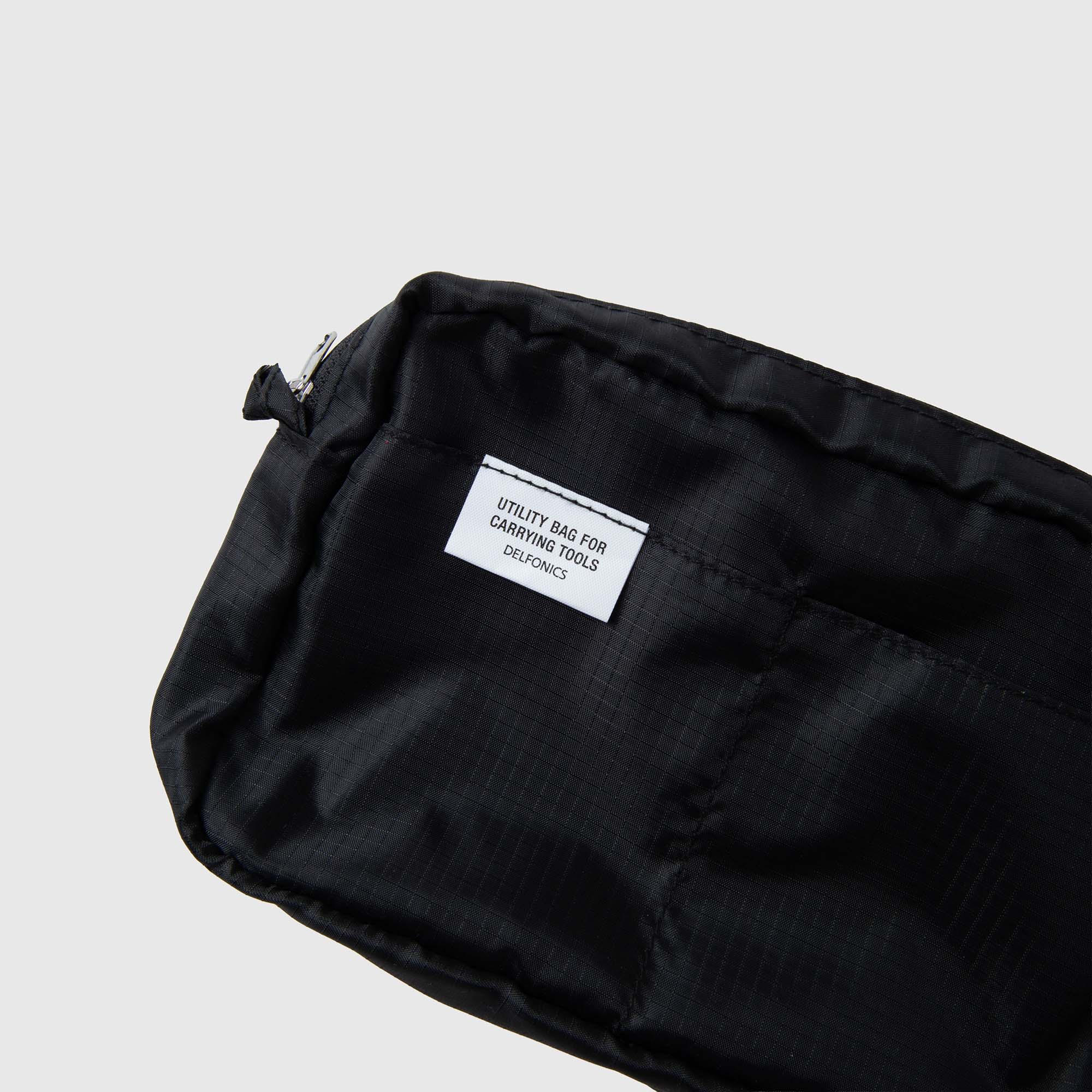 Eine schwarze Delfonics-Innentasche „AIR S“ aus strapazierfähigem Ripstop-Gewebe mit einem weißen Etikett, auf dem „UTILITY BAG FOR CARRYING TOOLS DELUXE“ steht, abgebildet vor einem einfarbigen hellgrauen Hintergrund. Die leichte Tasche von Delfonics hat die perfekte Größe, um wichtige Dinge wie Ihr Notizbuch im A6-Format und andere kleine Werkzeuge zu transportieren.