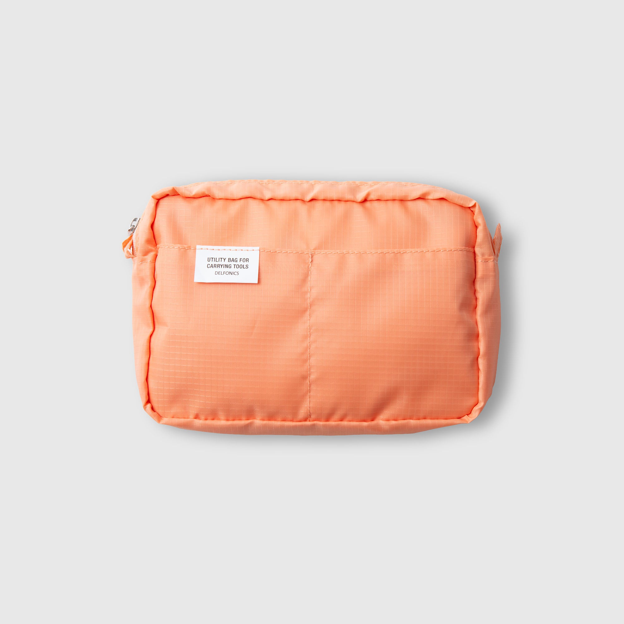 Eine kleine, rechteckige, orangefarbene Delfonics-Innentasche „AIR S“ aus strapazierfähigem Ripstop-Gewebe mit einer Vordertasche, auf der ein weißes Etikett mit der Aufschrift „Utility bag for daily use“ angebracht ist, ist vor einem schlichten hellgrauen Hintergrund abgebildet.