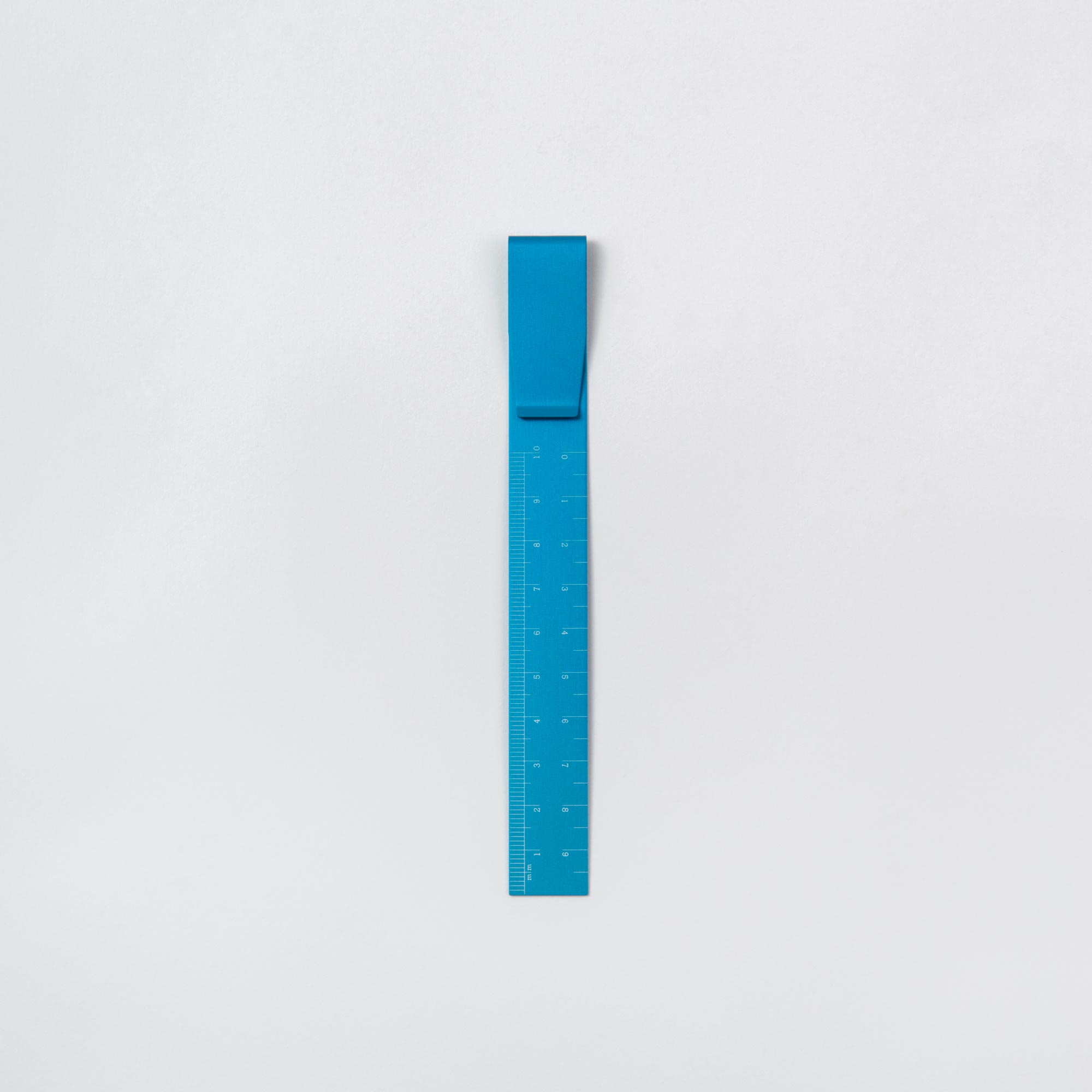 Das Hightide Clip Ruler – das Lineal To Go, ein blaues flexibles Lineal mit metrischen Maßangaben, steht vertikal auf hellgrauem Untergrund. Sein oberes Ende verfügt über eine breitere gefaltete Lasche, ideal für Bullet Journal-Fans, die Wert auf Präzision und Stil legen.