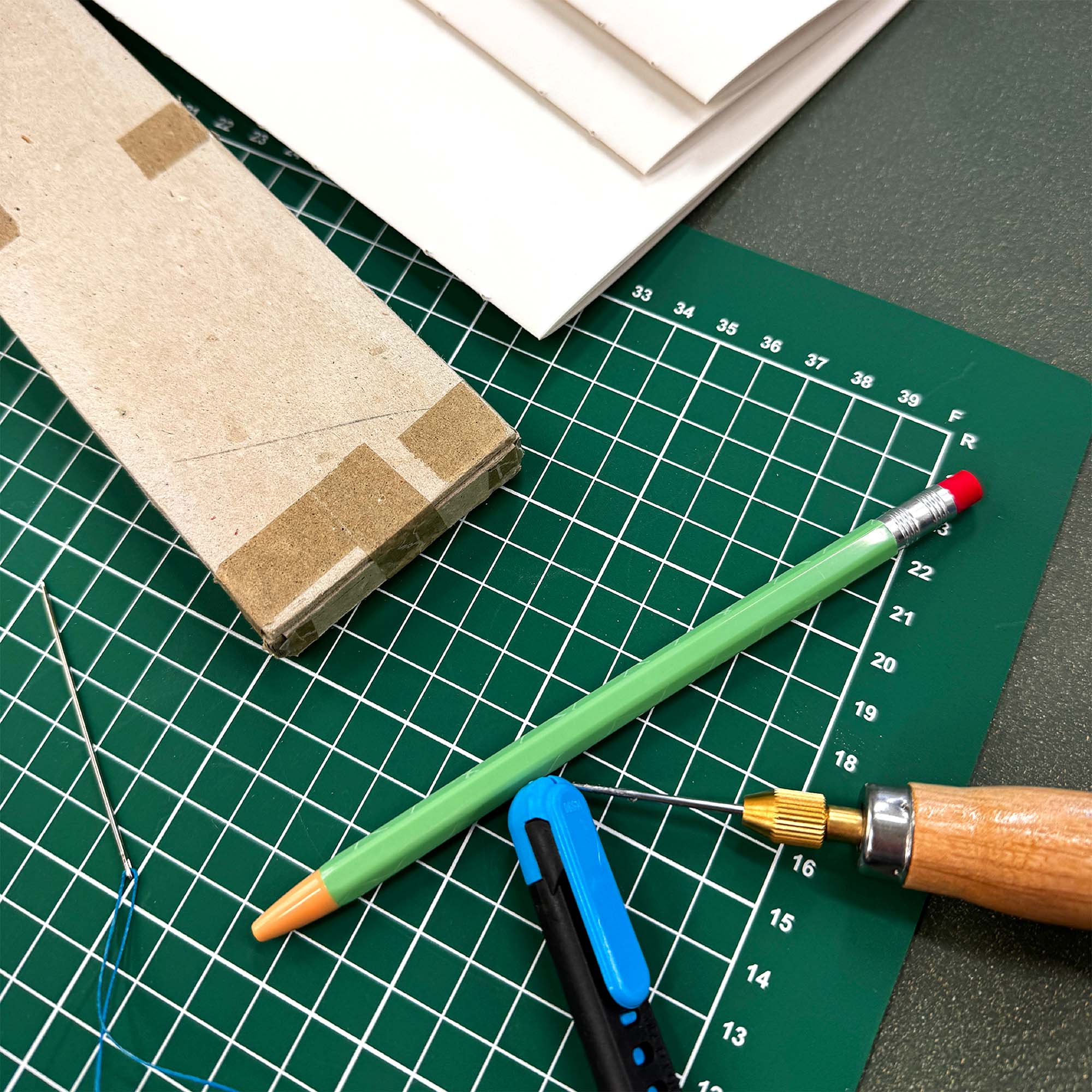Ein grüner Bleistift, ein blaues Universalmesser und ein Holzwerkzeug liegen auf einer Schneidematte mit Papier - ideales Zubehör für den Workshop Crémant und Notizbuch von paperlux.store, perfekt für die Buchbinderei mit Langstichheftung.