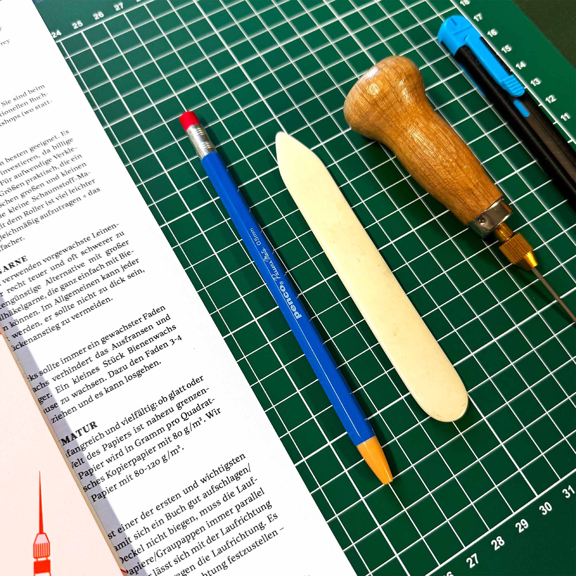 Auf einer grünen Schneidematte von paperlux.store liegen neben einem aufgeschlagenen Deutschbuch ein Bleistift, ein Falzbein, eine Ahle und ein Universalmesser - wichtige Werkzeuge für Ihren Workshop Crémant und Notizbuch.