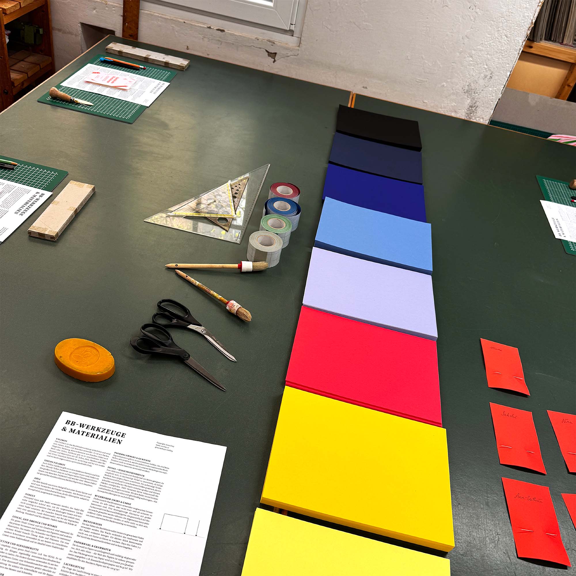Ein Tisch für den Workshop Crémant und Notizbuch von paperlux.store mit bunten rechteckigen Papieren, Scheren, Klebeband, Linealen, einem Pinsel und gedruckten Anleitungen, die um den Arbeitsbereich herum angeordnet sind.