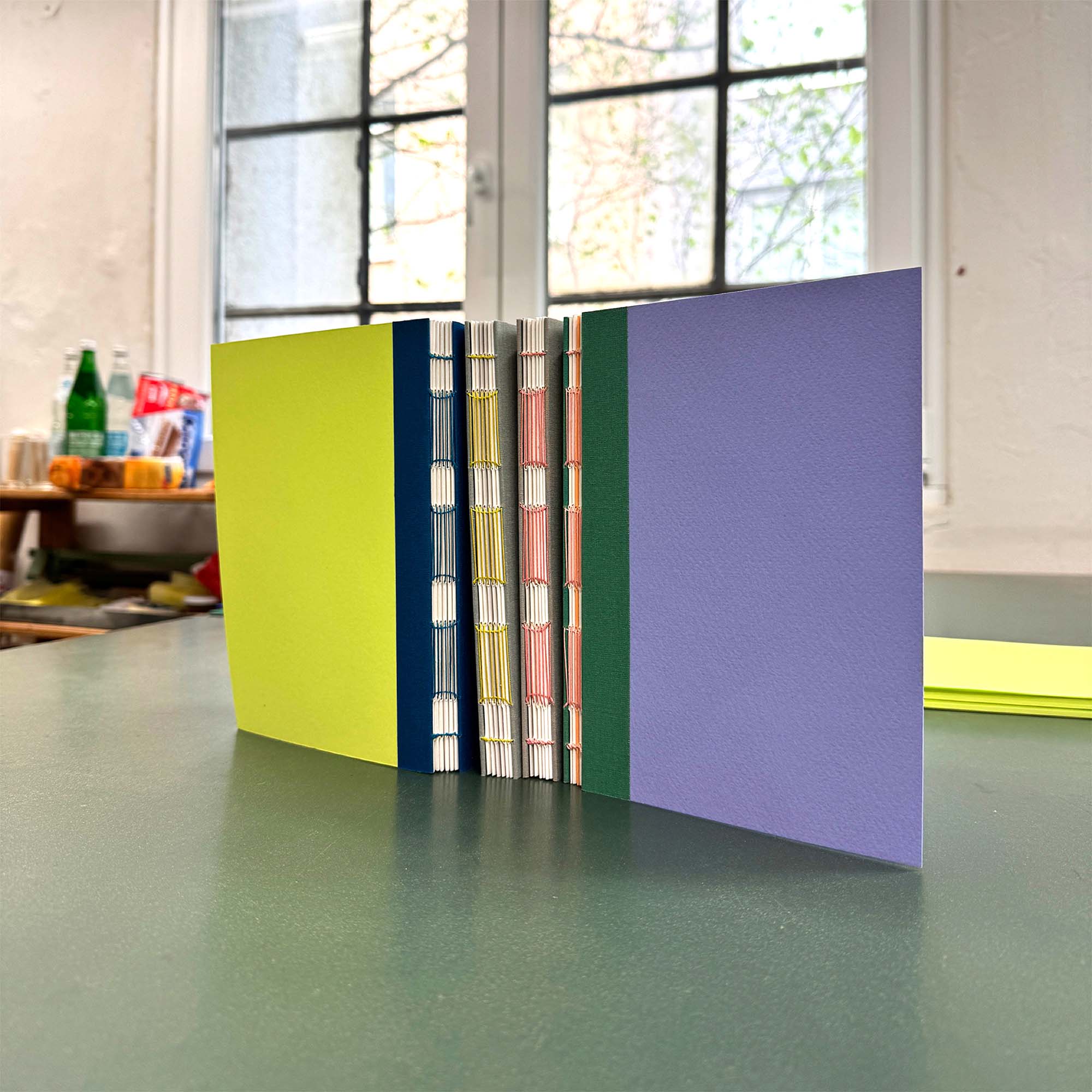 Ein handgefertigtes Notizbuch aus dem Workshop Crémant und Notizbuch von paperlux.store mit gelb-grünem und lila Einband, Langstichheftung und sichtbaren Fäden steht aufrecht auf einem grünen Tisch; Fenster und Regale im Hintergrund.
