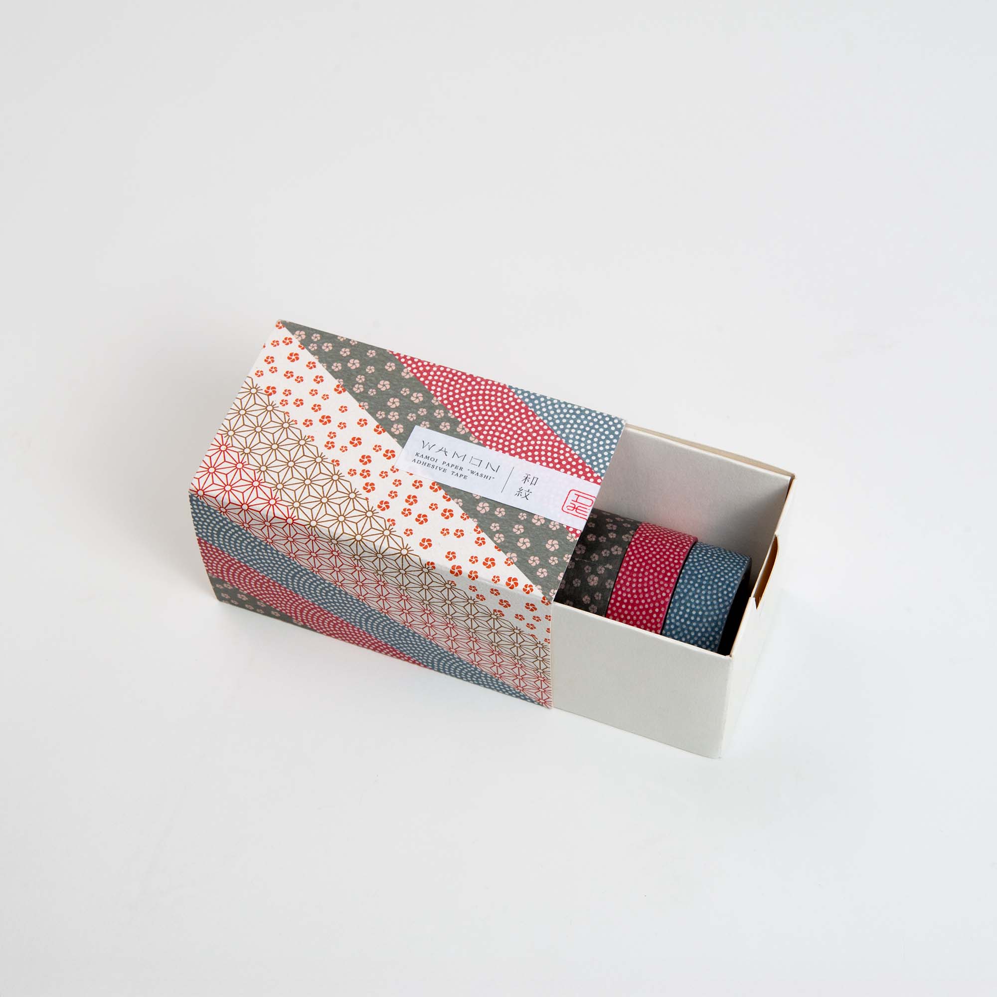 Die Kamoi Kakoshi mt Washi Tape - Wamon 5 Geschenkbox mit farbenfrohem, geometrischem Design ist auf einer weißen Oberfläche teilweise geöffnet und zeigt drei Rollen Washi Tape in Blau, Rot und Grau mit japanischen Mustern im Inneren.