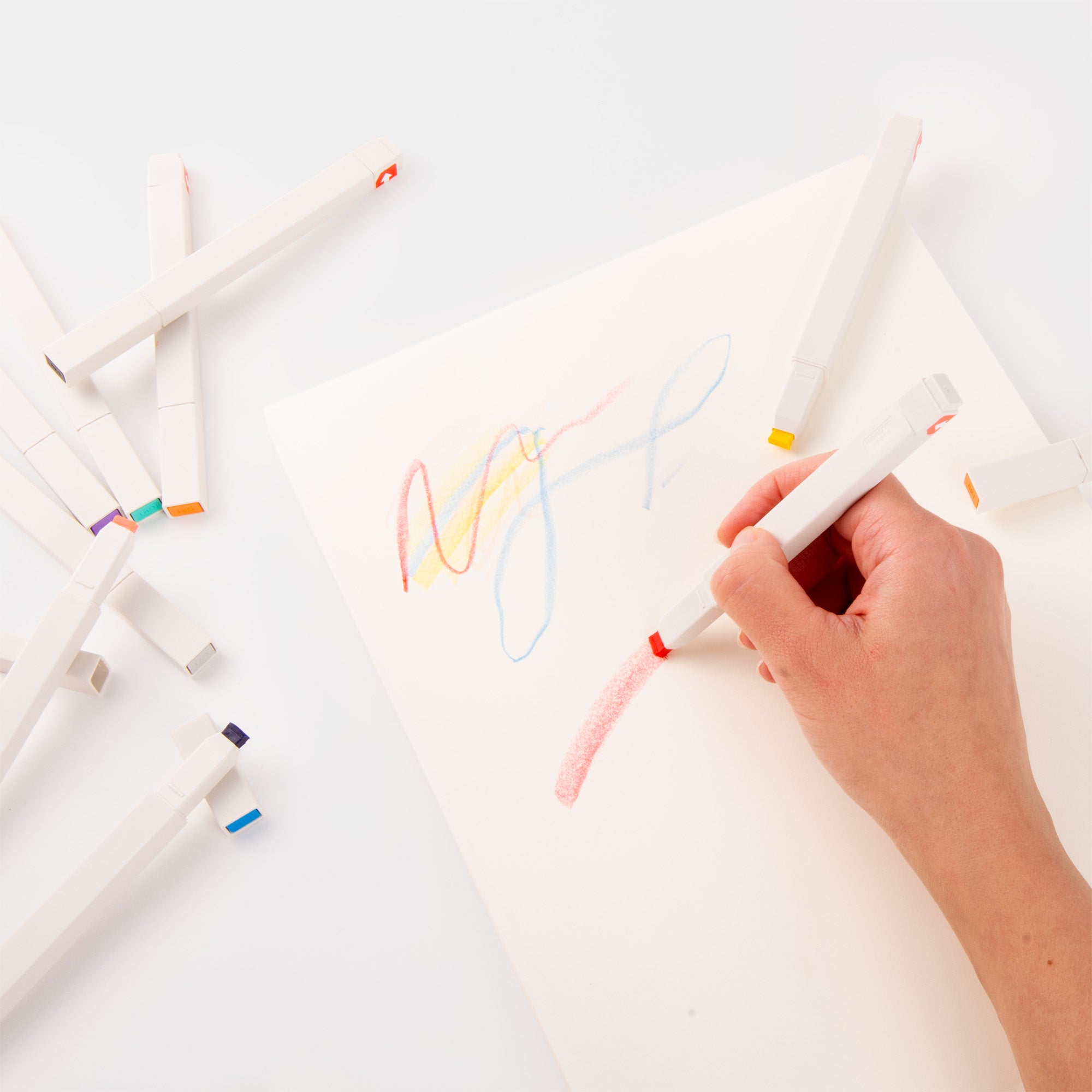 Eine Hand zeichnet mit einem roten Kokuyo Pasta Marker aus dem Set 10 Farben auf weißem Papier, während andere Marker aus dem Set mit Pastellfarben-Spitzen auf einer weißen Fläche verstreut sind.