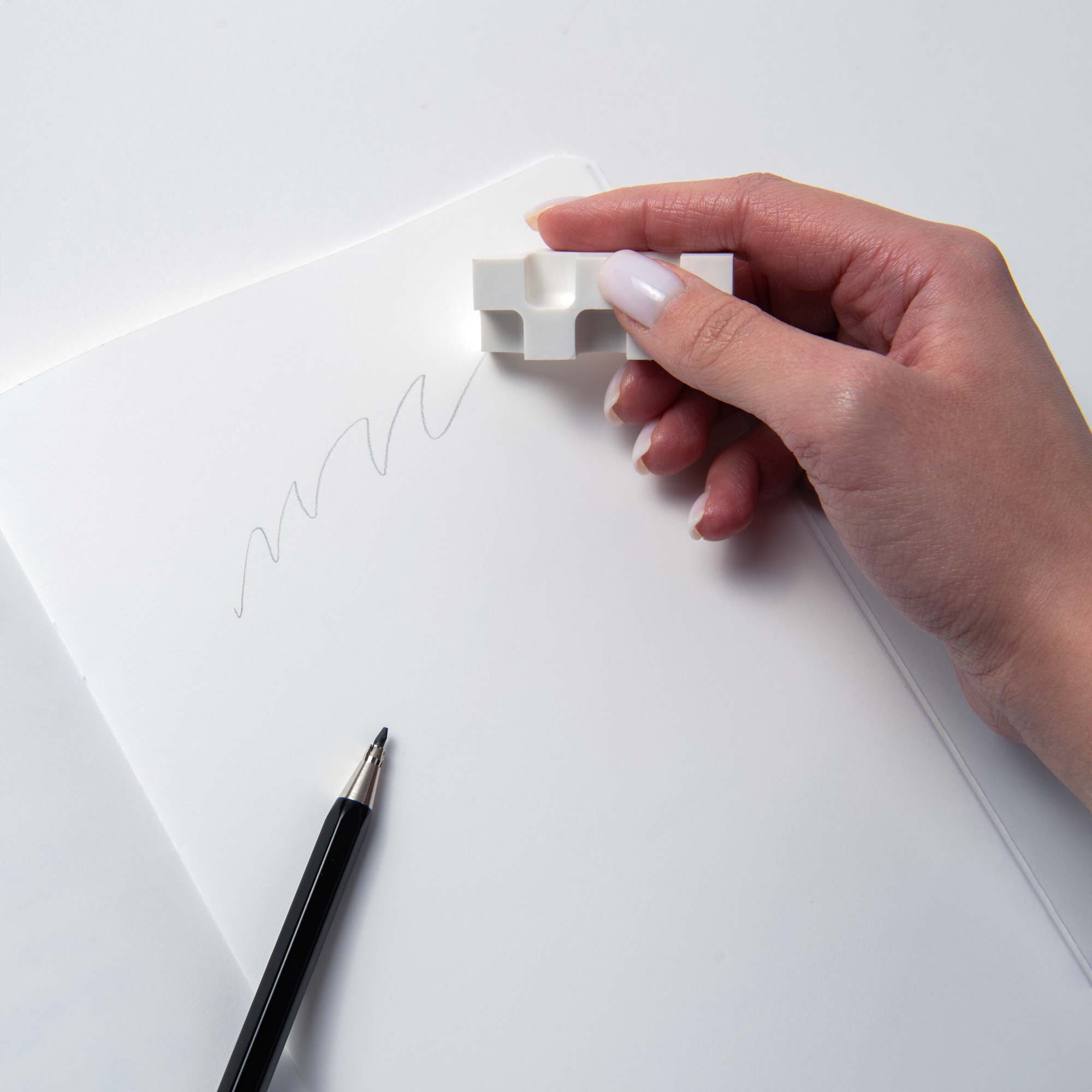 Eine Hand verwendet einen puzzleförmigen Kokuyo-Radiergummi „Würfel“, um einen verschnörkelten Bleistiftstrich auf weißem Papier auszuradieren. Ein schwarzer Bleistift liegt daneben auf dem Schreibtisch. Der Radiergummi weist ineinander greifende Teile auf.