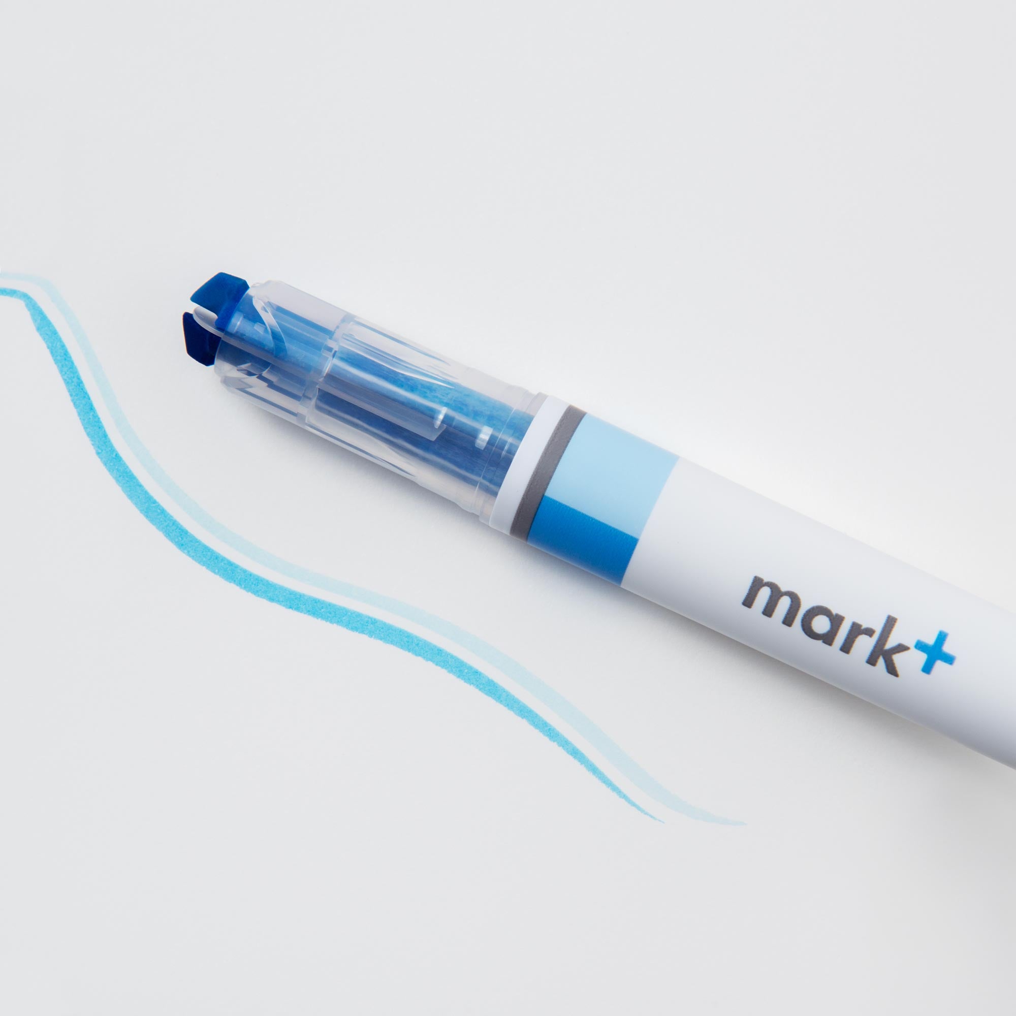 Ein Kokuyo Ein (Text-) Marker, Zwei Farben mit einer kräftigen blauen Meißelspitze und der Aufschrift „mark+“ auf seinem Körper liegt auf einer weißen Oberfläche neben zwei von ihm erzeugten blauen Wellenlinien.