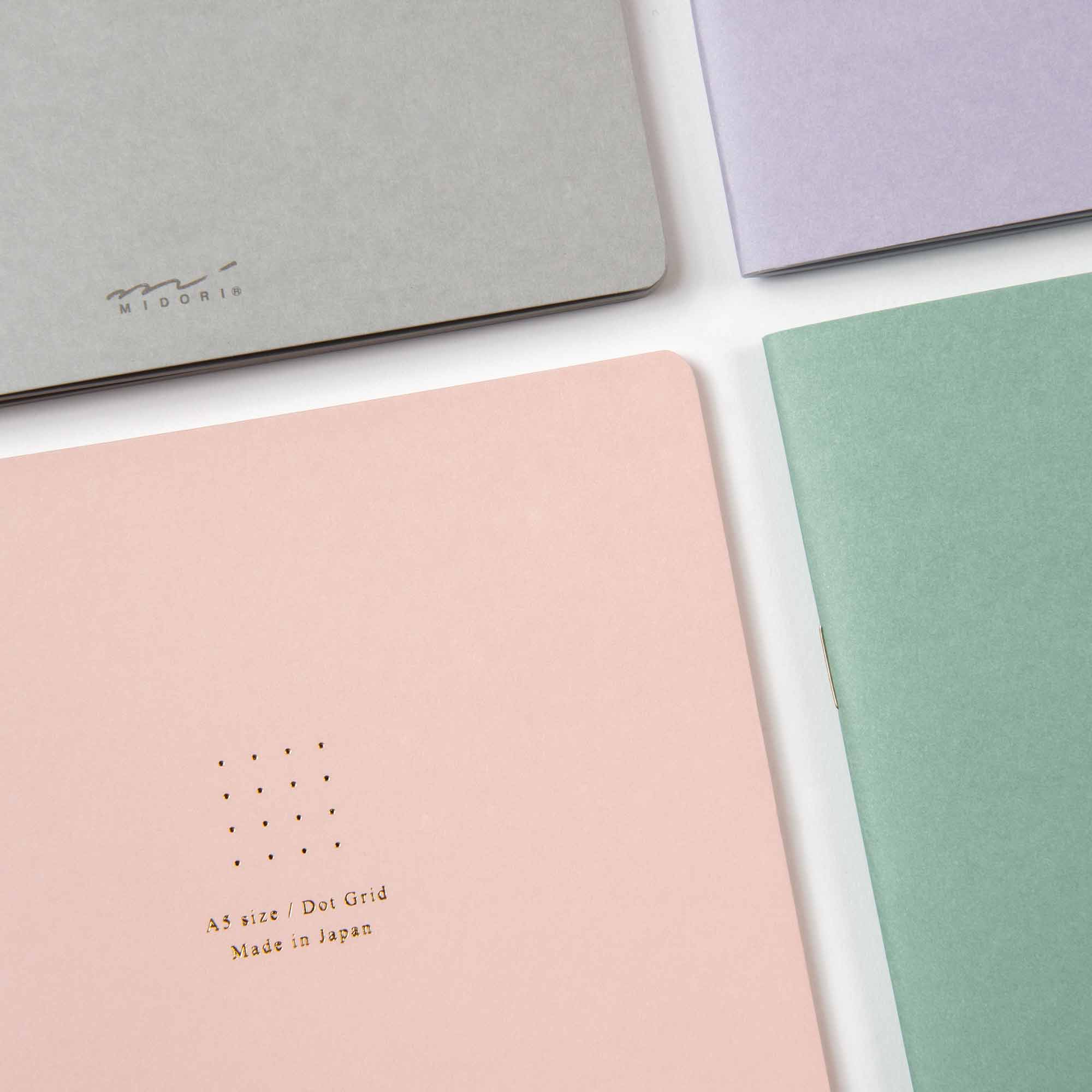 Midori Notizhefte Color Dot Grid werden auf einer weißen Oberfläche mit Umschlägen in Pastellrosa, Grün, Grau und Lavendel präsentiert. Das rosafarbene hat ein gepunktetes Gitterdesign mit dem Text „A5-Größe / Dot Grid Made in Japan“.