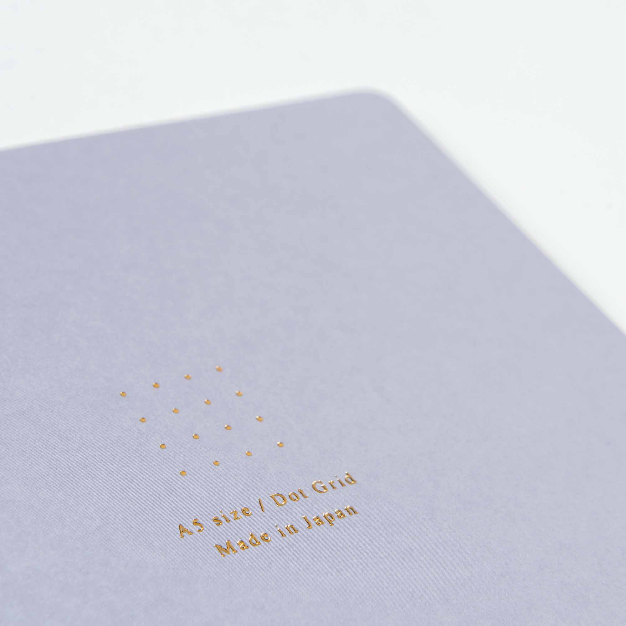 Nahaufnahme eines hellgrauen Midori-Notizhefte Color Dot Grid-Notizbuchumschlags mit goldgeprägtem Text „A5-Größe / Dot Grid Made in Japan“ und sichtbarem Punktrastermuster, das die handwerkliche Heißfolienprägung zur Schau stellt.