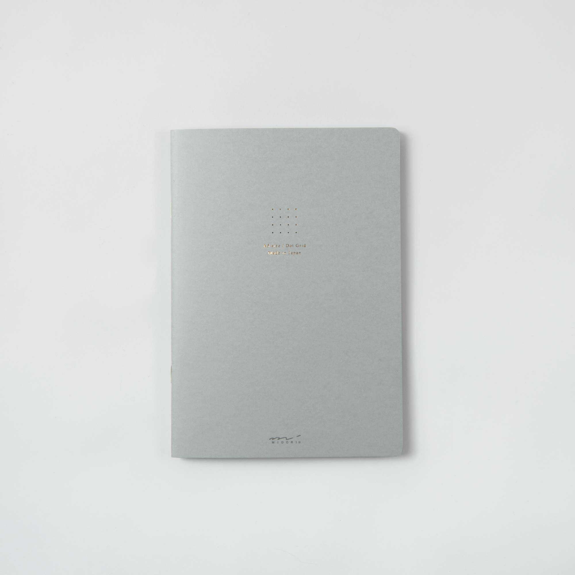 Ein minimalistisches, hellgraues Midori-Notizheft „Color Dot Grid“ mit heißfoliengeprägtem Goldtext in der Mitte nahe der Oberseite ruht auf einer weißen Oberfläche und verleiht Ihrem Arbeitsplatz einen Hauch von Eleganz.