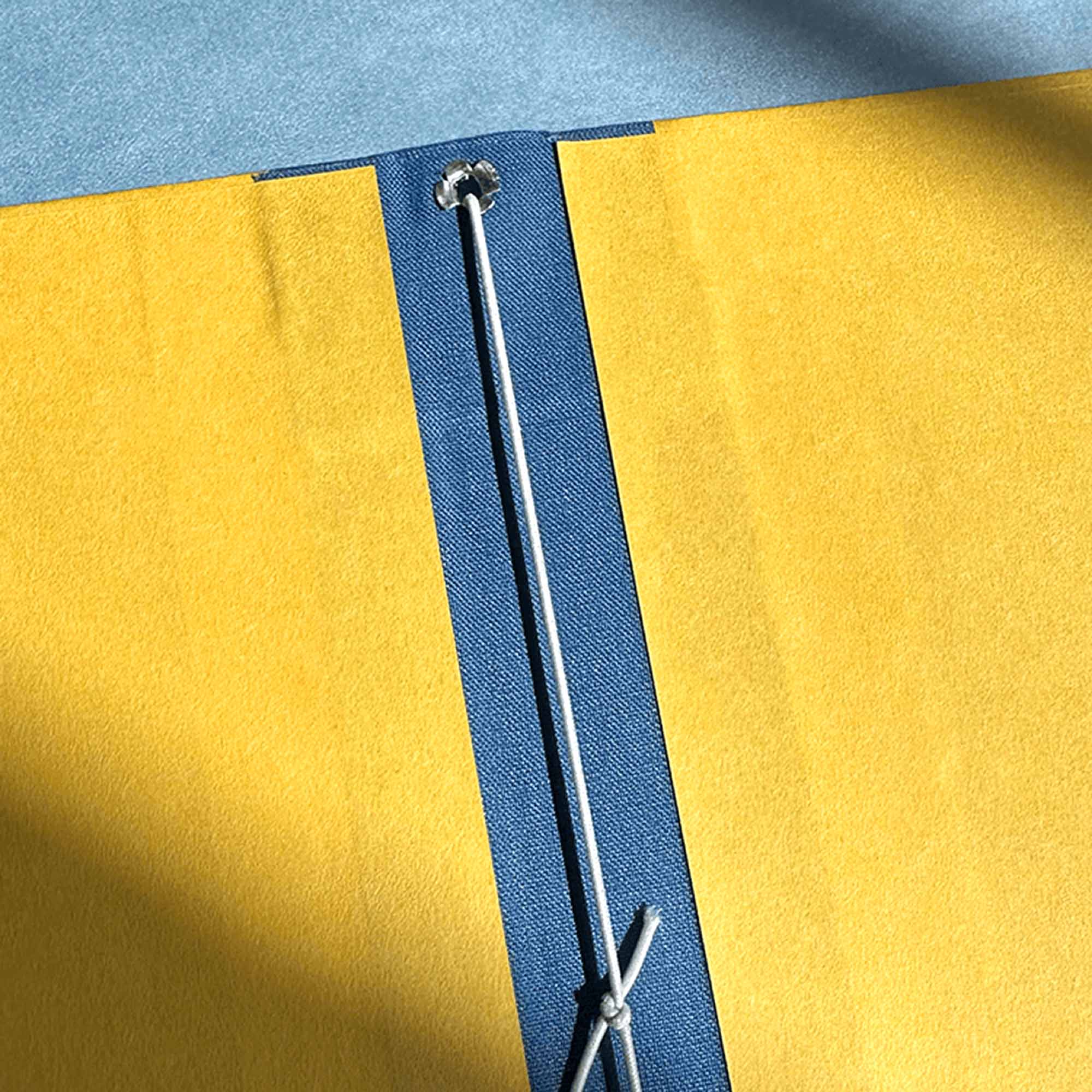 Eine Nahaufnahme des Workshops Modulares Notizbuch von paperlux.store: eine gelbe Mappe mit blauem Rücken, weiße Schnurbindung durch metallverstärkte Löcher, Sonnenlicht hebt die strukturierte Oberfläche hervor.