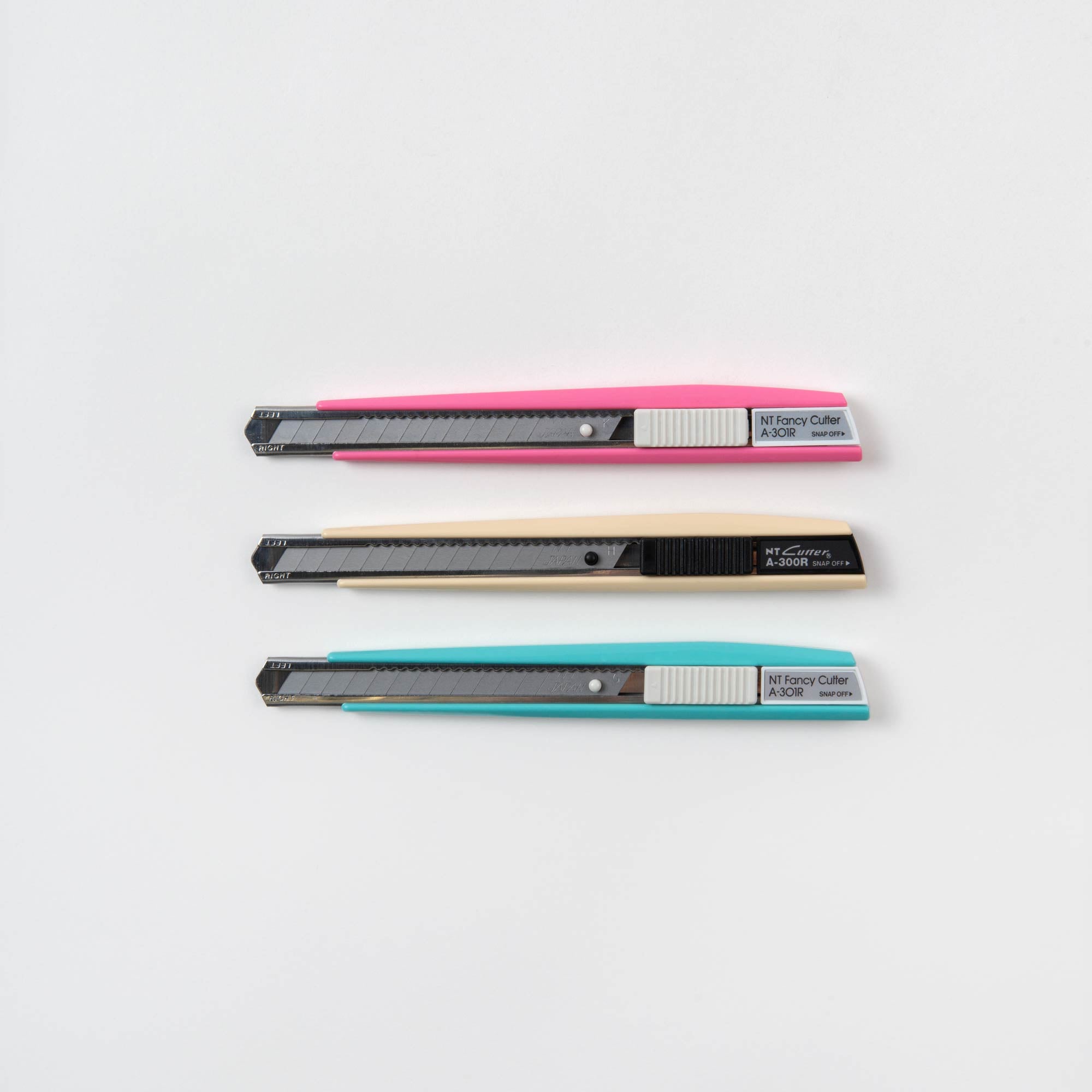 Drei einziehbare Universalschneider „Cutter in Colour“ von NT Cutter auf weißem Hintergrund mit silbernen Klingen und Schiebemechanismus. Die Griffe sind rosa (oben), schwarz (Mitte) und hellblau (unten), ideal zum einfachen Schneiden bei Bastelprojekten.