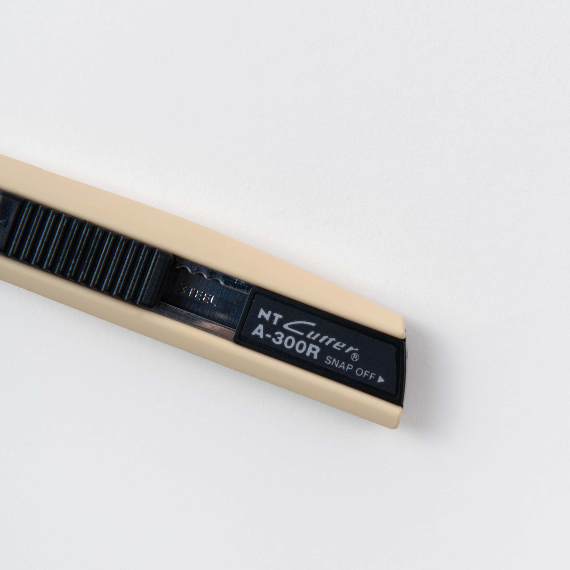 Nahaufnahme eines beigen NT Cutter Universalmessers mit schwarzem geriffeltem Griff, teilweise ausgefahrener Klinge und dem Aufdruck „Cutter in Colour“ am Ende. Ideal für präzise Schneidearbeiten oder Bastelprojekte, liegt es auf einer weißen Oberfläche.