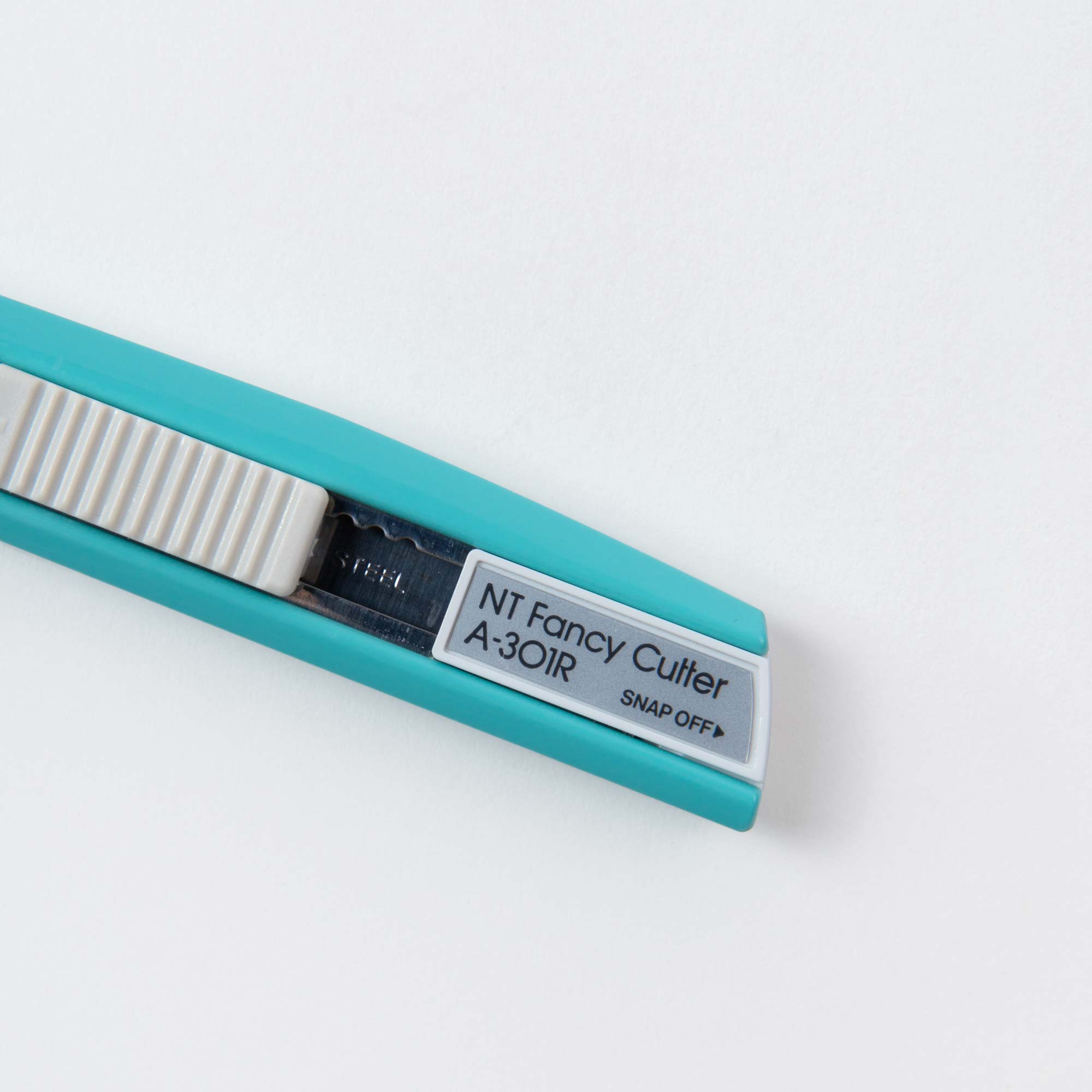 Ein blaugrünes NT Cutter-Universalmesser namens „Cutter in Colour“ verfügt über einen weißen Schieber und eine sichtbare scharfe Klinge mit „SNAP OFF“, ideal für präzises Schneiden und Basteln, vor einem weißen Hintergrund.