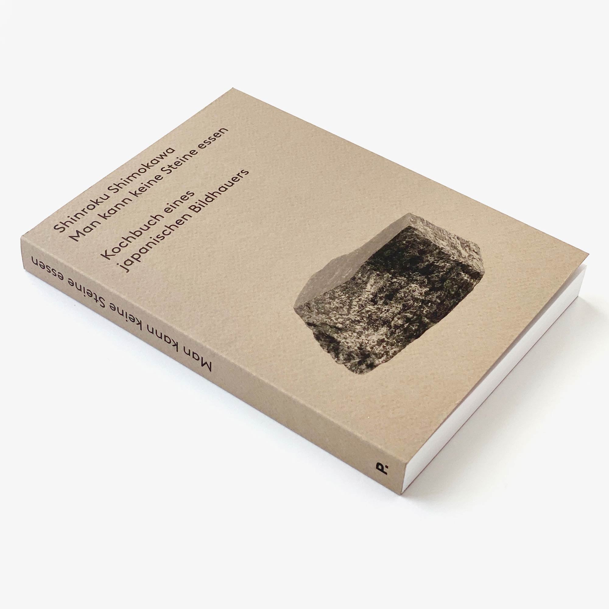 Das Taschenbuch „Man kann keine Steine essen: Kochbuch eines japanischen Bildhauers“ von Shinroku Shimokawa von Prima Publikationen liegt auf einer weißen Unterlage. Sein mit einem grauen Steinbild geschmückter Einband ähnelt in seiner Erkundung der kulinarischen Tiefe auf faszinierende Weise den selbstgefertigten Kochutensilien.