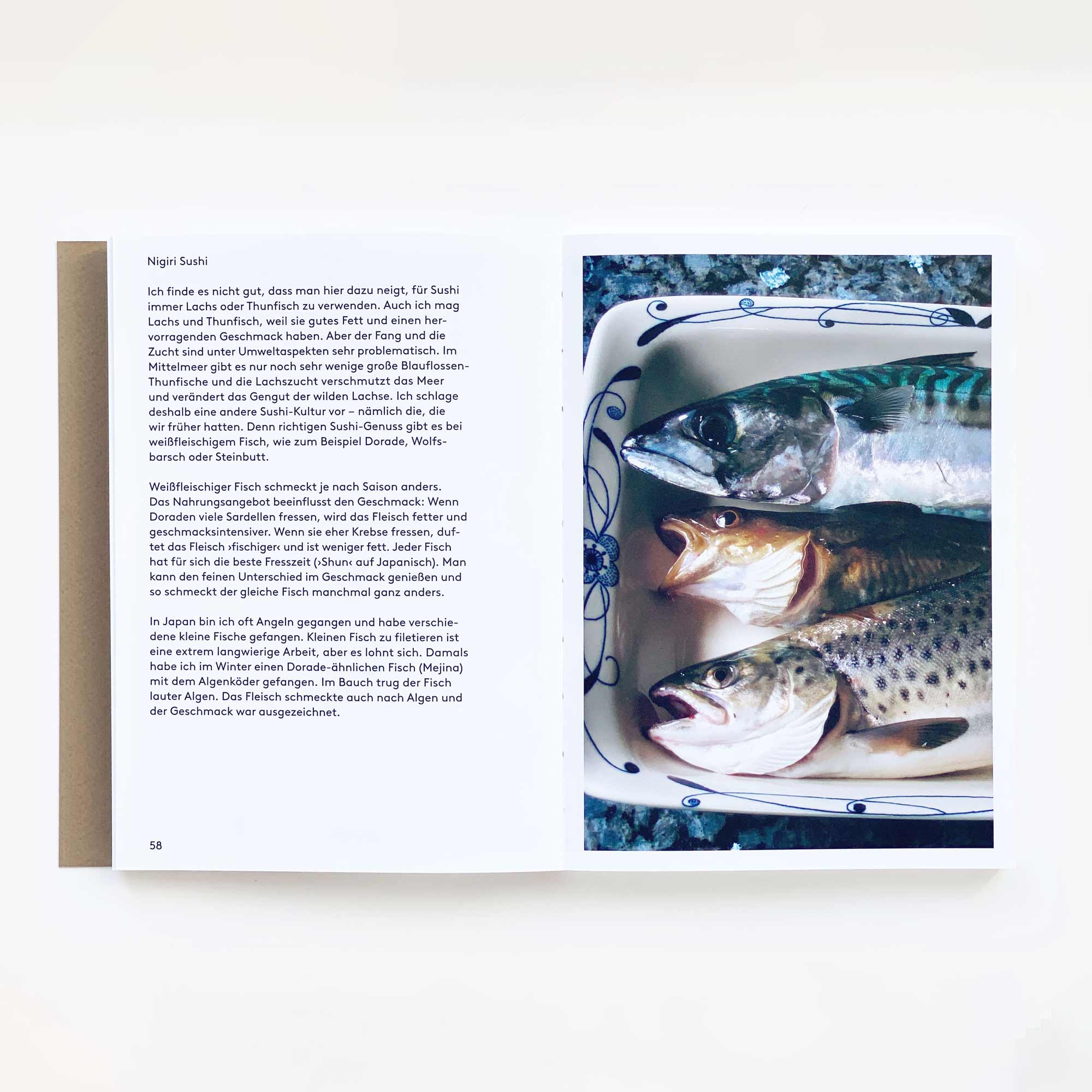 Das aufgeschlagene Buch zeigt rechts ein Bild von drei ganzen Fischen in einer gemusterten Schale und links einen deutschen Text über Japanische Küche und Sushi. Das Shinroku Shimokawa-Kochbuch von Prima Publikationen ist auf einer weißen Fläche abgebildet.
