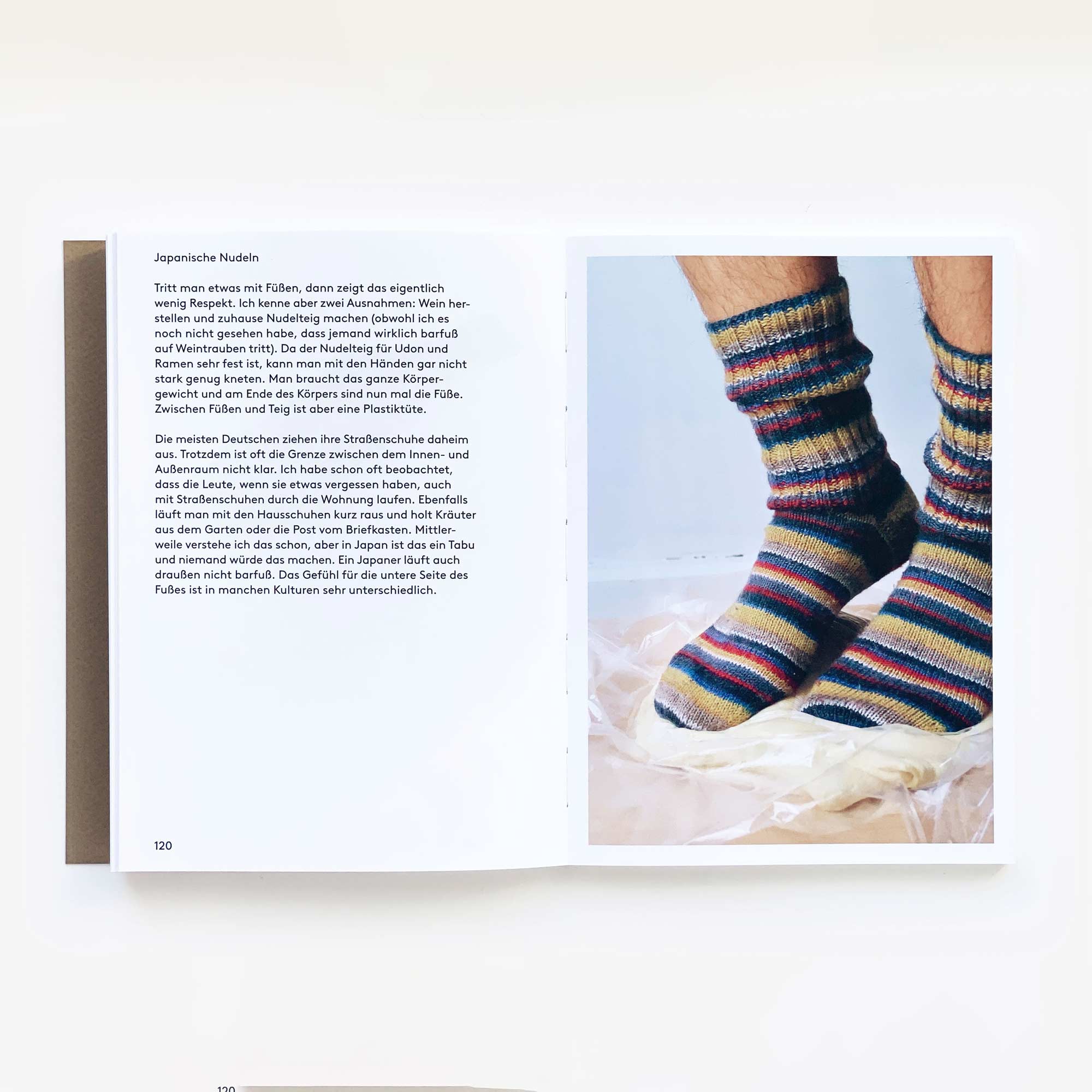 In „Man kann keine Steine essen“ von Shinroku Shimokawa zeigt ein aufgeschlagenes Buch deutschen Text und ein Bild von gestreiften Stricksocken auf Stoff. Der neutrale Hintergrund wertet die Socken auf, so wie selbstgefertigte Kochutensilien authentische Details hervorheben. Erhältlich bei Prima Publikationen.