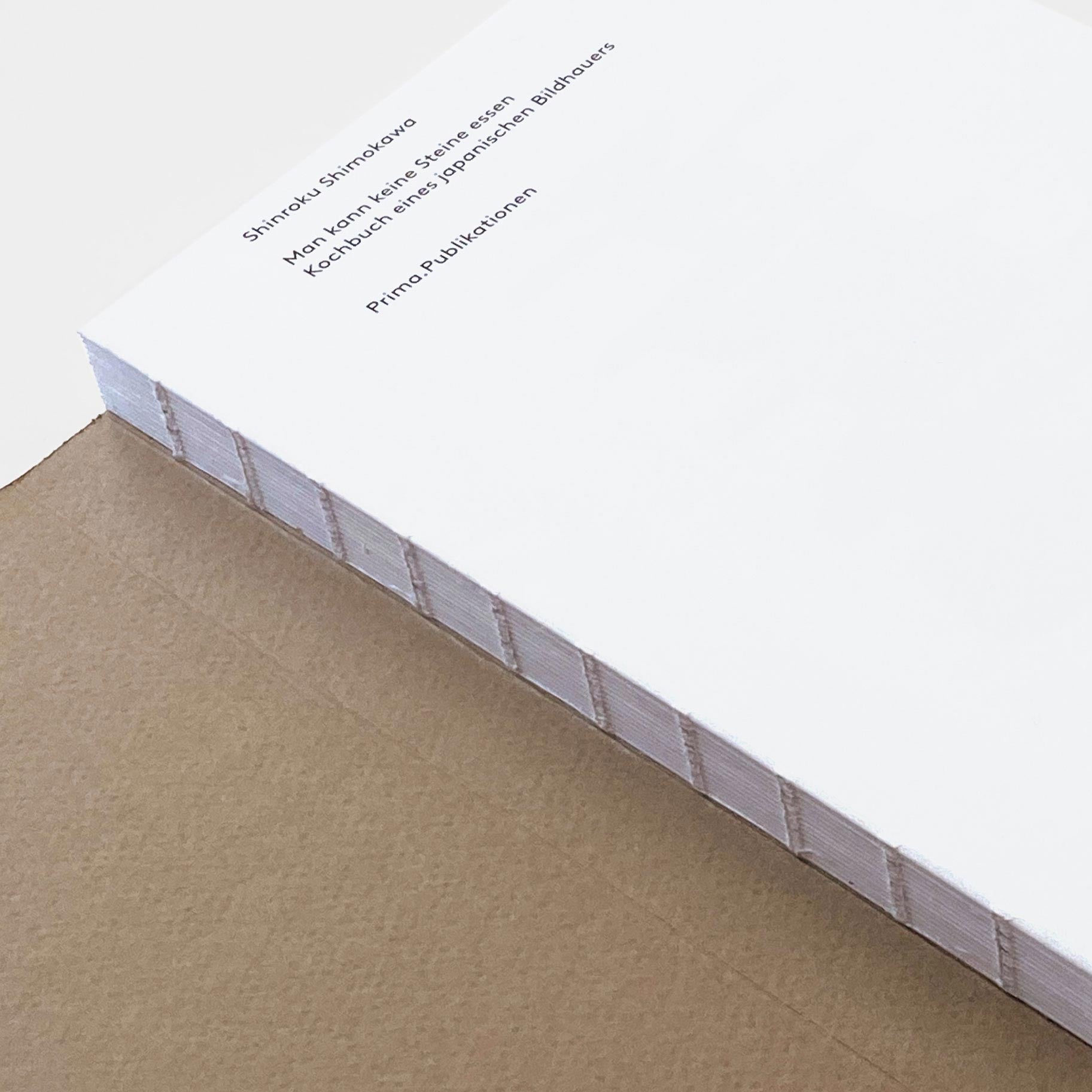 Eine Nahaufnahme eines Stapels weißes Druckerpapier mit kleinem, meist unleserlichem Text auf dem obersten Blatt. Es weist auf „Shinroku Shimokawa – Man kann keine Steine essen: Kochbuch eines japanischen Bildhauers“ von Prima Publikationen hin und listet möglicherweise regionale Zutaten oder handgefertigte Utensilien auf.