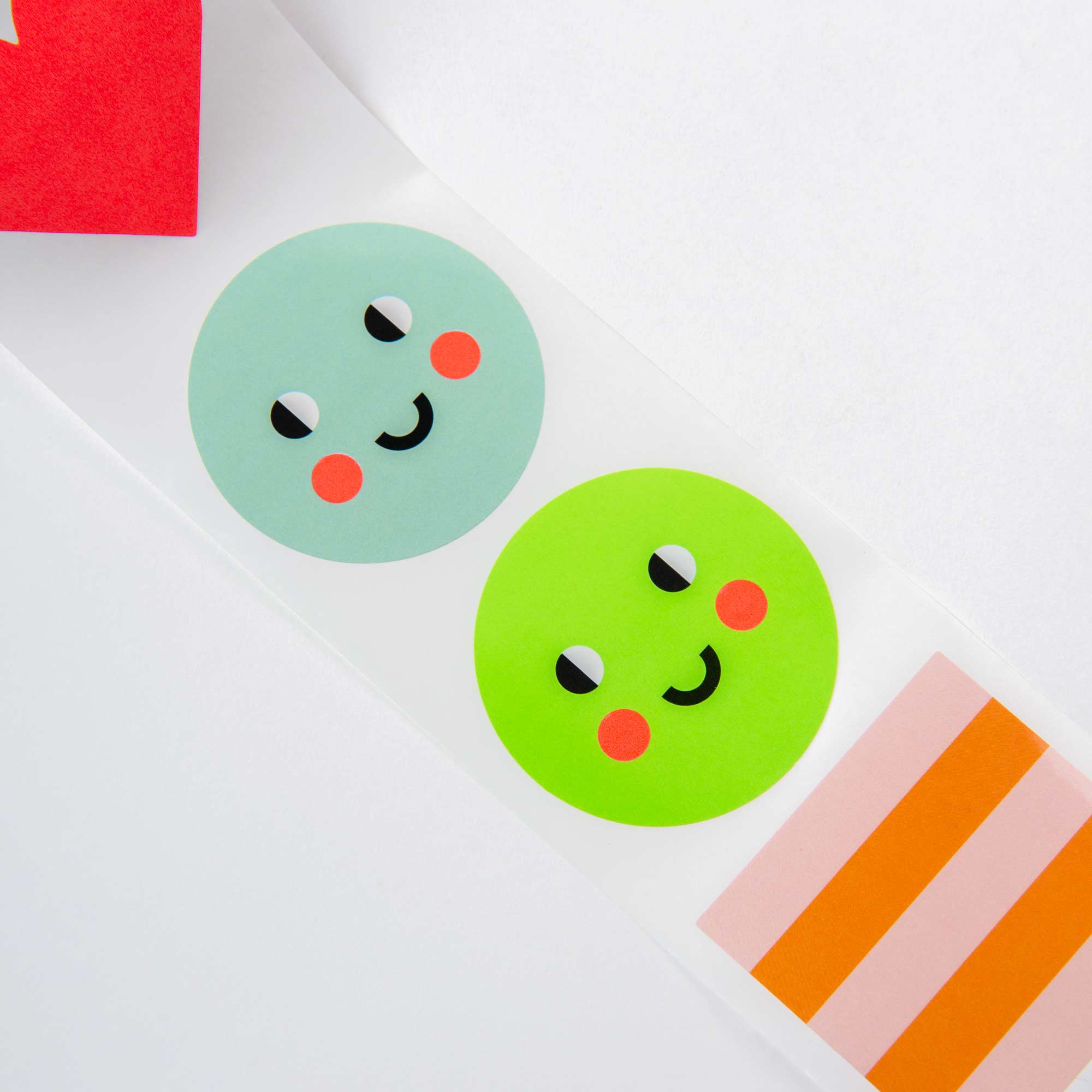 Das Sticker-Set - 7 Mal Fröhlichkeit von Paper Poetry enthält eine verspielte Auswahl farbenfroher Sticker mit zwei fröhlichen Smileys in Blau und Grün mit rosigen Wangen, ideal für Geburtstagsfeiern. Es enthält rosa und orange gestreifte Blöcke sowie ein teilweise sichtbares kleines rotes Dreieck.
