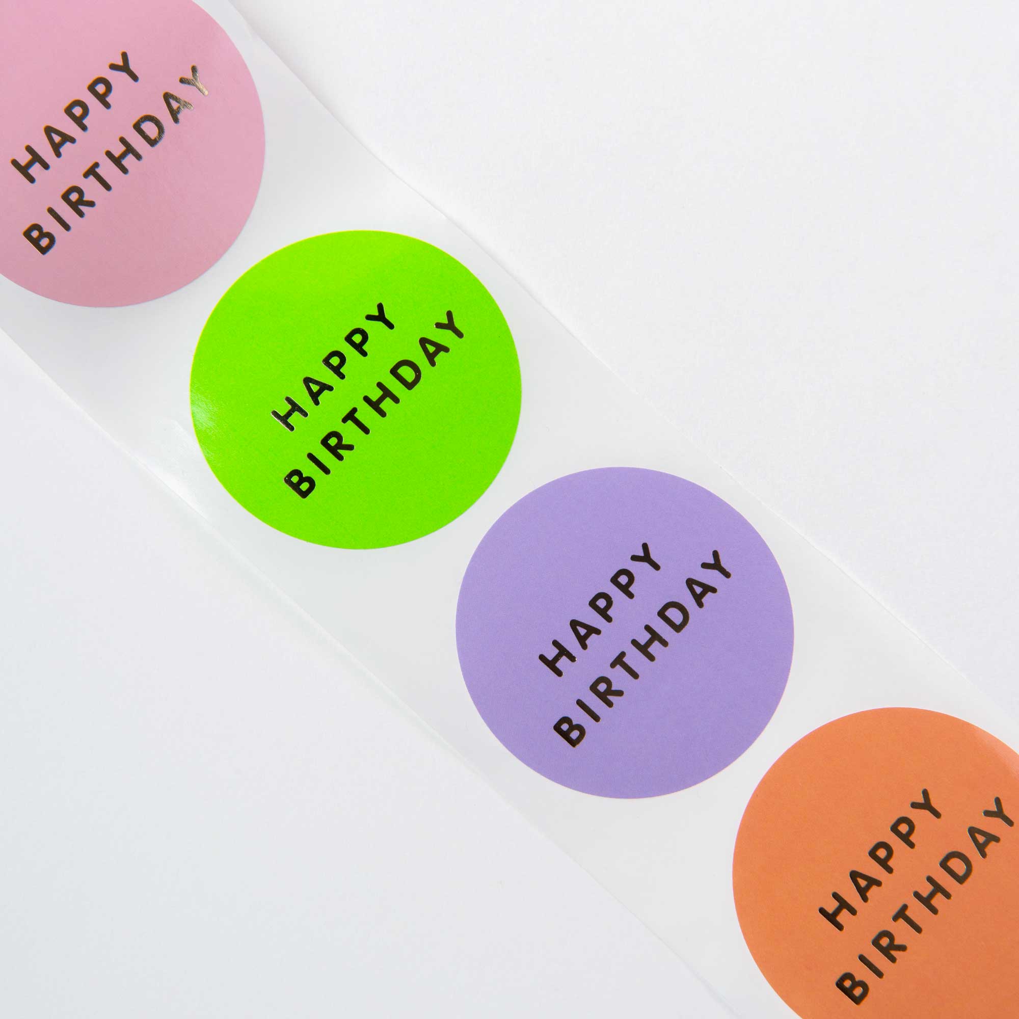 Ein Streifen mit vier runden „Happy Happy Birthday!“-Aufklebern von Paper Poetry in leuchtenden Rosa-, Grün-, Lila- und Orangetönen sind vertikal auf einem strahlend weißen Hintergrund angeordnet.