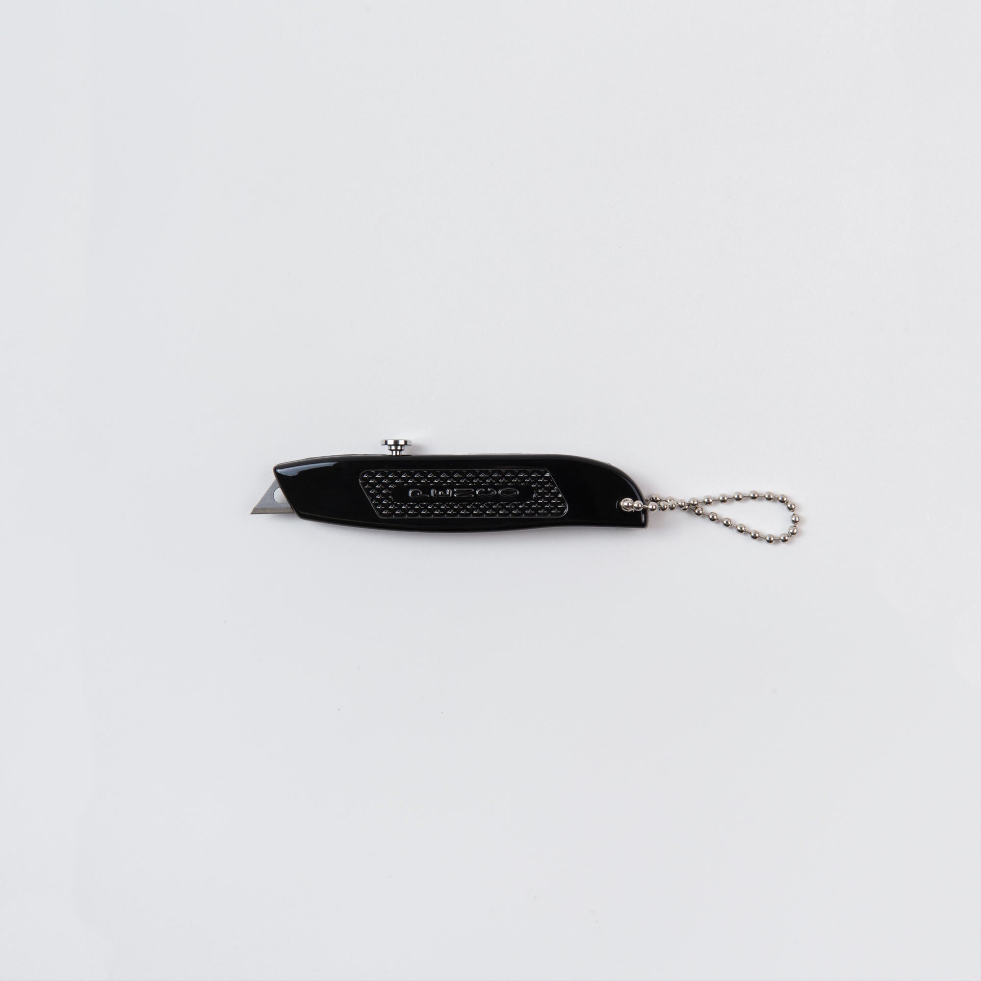 Das Allzweckmesser oder „der Cutter an der Kette“ von Penco ist ein kompaktes schwarzes Universalmesser mit einziehbarer Klinge und strukturiertem Griff. Es verfügt über eine silberne Kette zum bequemen Aufhängen und sieht elegant vor einem schlichten weißen Hintergrund aus.