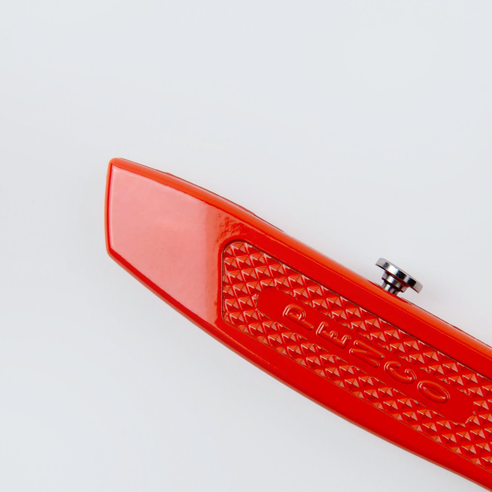 Nahaufnahme des Penco Allzweckmessers - der Cutter an der Kette - mit roter, strukturierter, dreieckig gemusterter Oberfläche, abgerundeter Kante und kleinem Metallknopf auf der Oberseite; ideal zum Öffnen von Verpackungen und auf weißem Hintergrund fotografiert.
