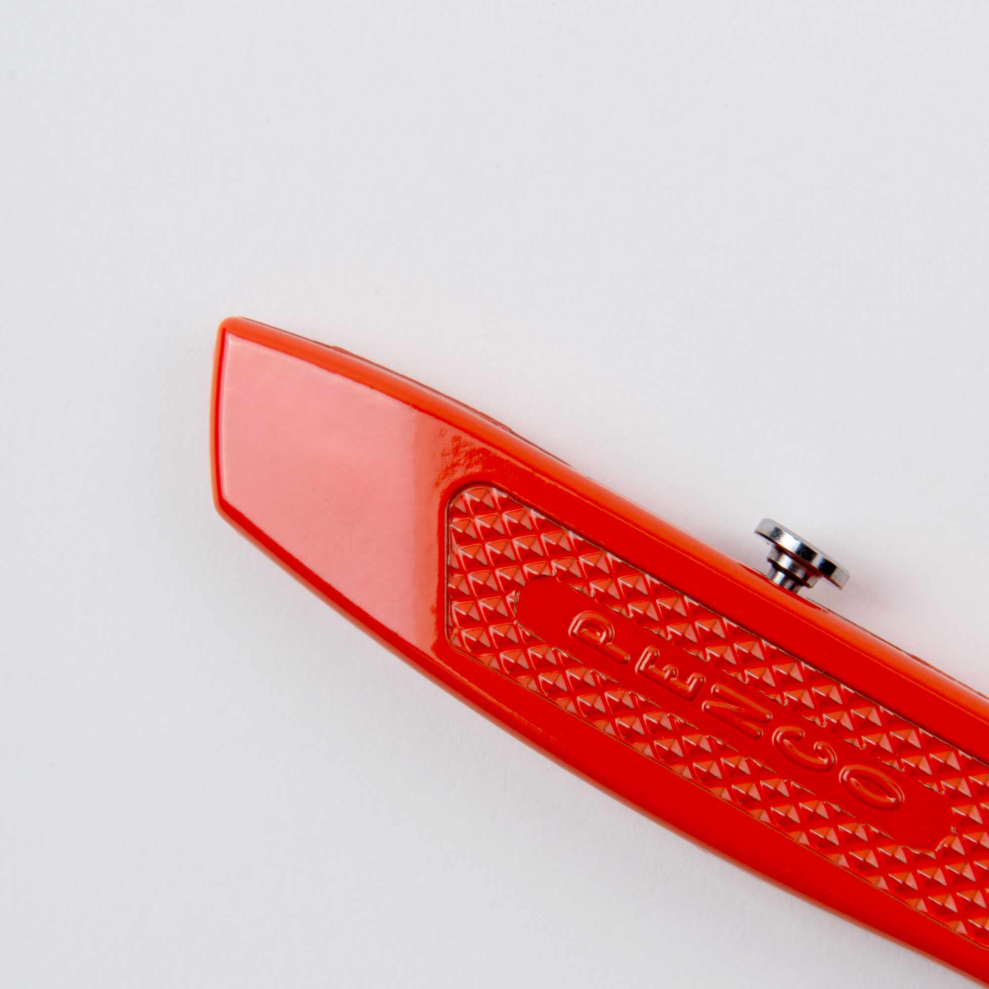 Nahaufnahme eines roten Plastikobjekts mit strukturierter Oberfläche mit der Aufschrift „PIESCO“ und einem kleinen silbernen Knopf, der darauf hinweist, dass es sich um Pencos Allzweckmesser handelt – oder auch: der Cutter an der Kette. Vor einem schlichten weißen Hintergrund.