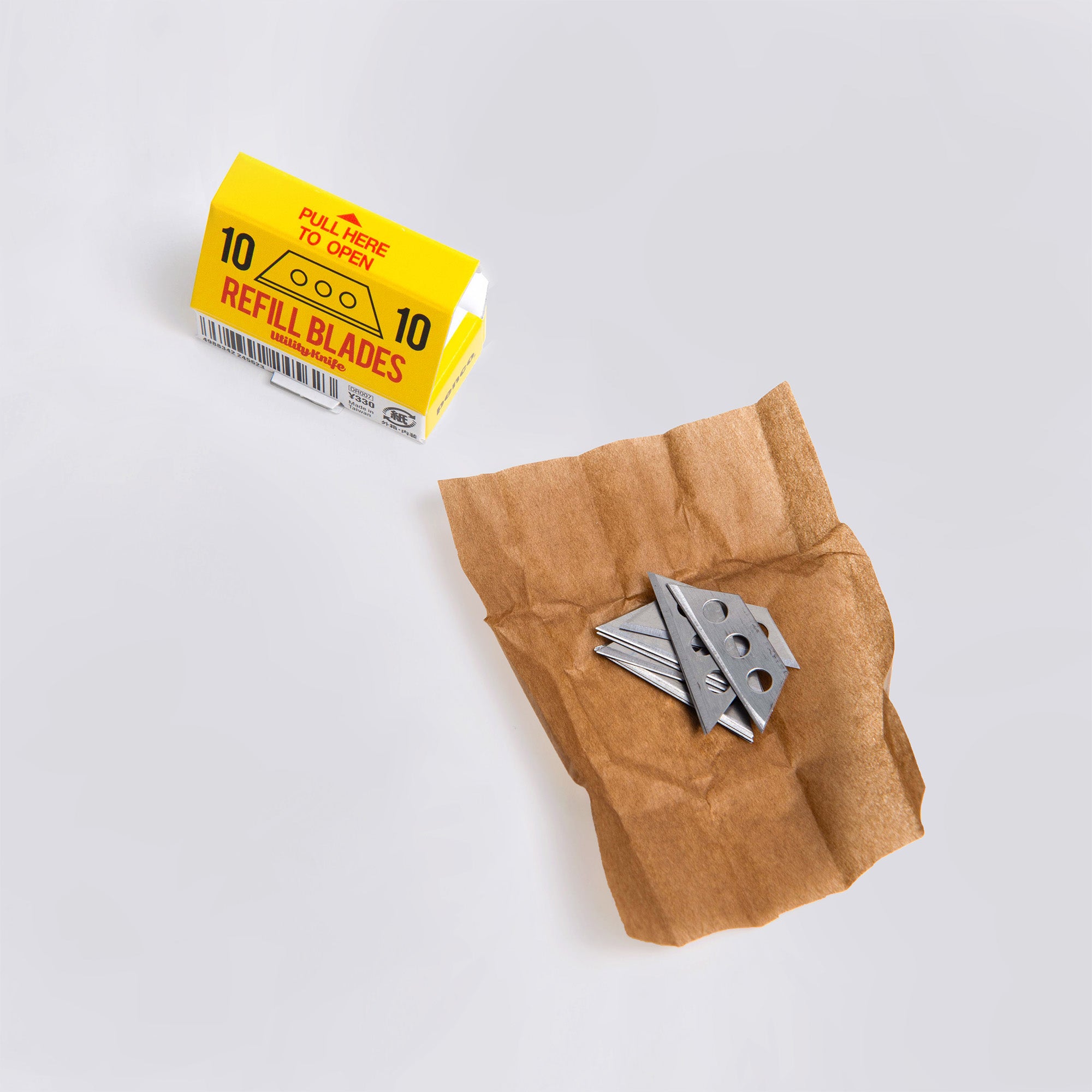 Eine kleine, geöffnete Packung mit Pencos „Ersatzklingen: Cutter an der Kette“ liegt neben zerknülltem braunem Papier. Auf dem Papier liegen zehn teilweise ausgepackte Edelstahlklingen. Die gelb-weiße Packung mit fettem Text ähnelt einem eleganten Allzweckmesser-Design.