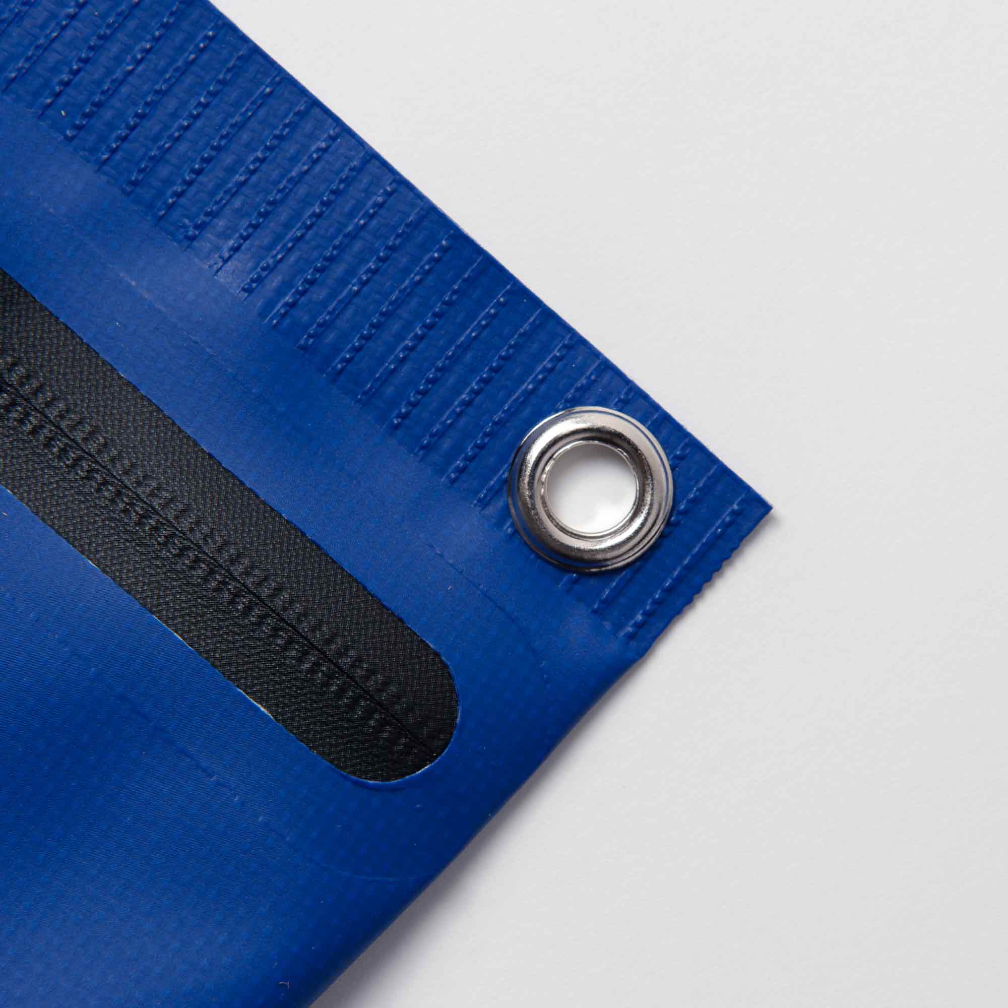 Nahaufnahme einer blauen Penco Tarp Pouch S mit Metallöse und strapazierfähigem Planenmaterial. Ein schwarzer verstärkter Streifen verläuft parallel zur Eckkante, mit strukturierter Naht in Randnähe. Der Hintergrund ist schlicht weiß.