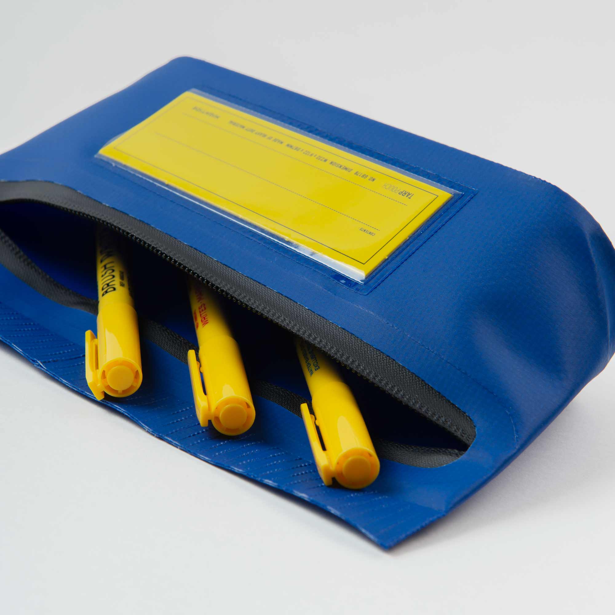 Die Penco Tarp Pouch S mit transparentem Etikettenhalter und wasserdichten Verschlüssen lässt sich öffnen und gibt mehrere gelbe Stifte preis. Das blaue Federmäppchen liegt auf einer schlichten weißen Oberfläche.