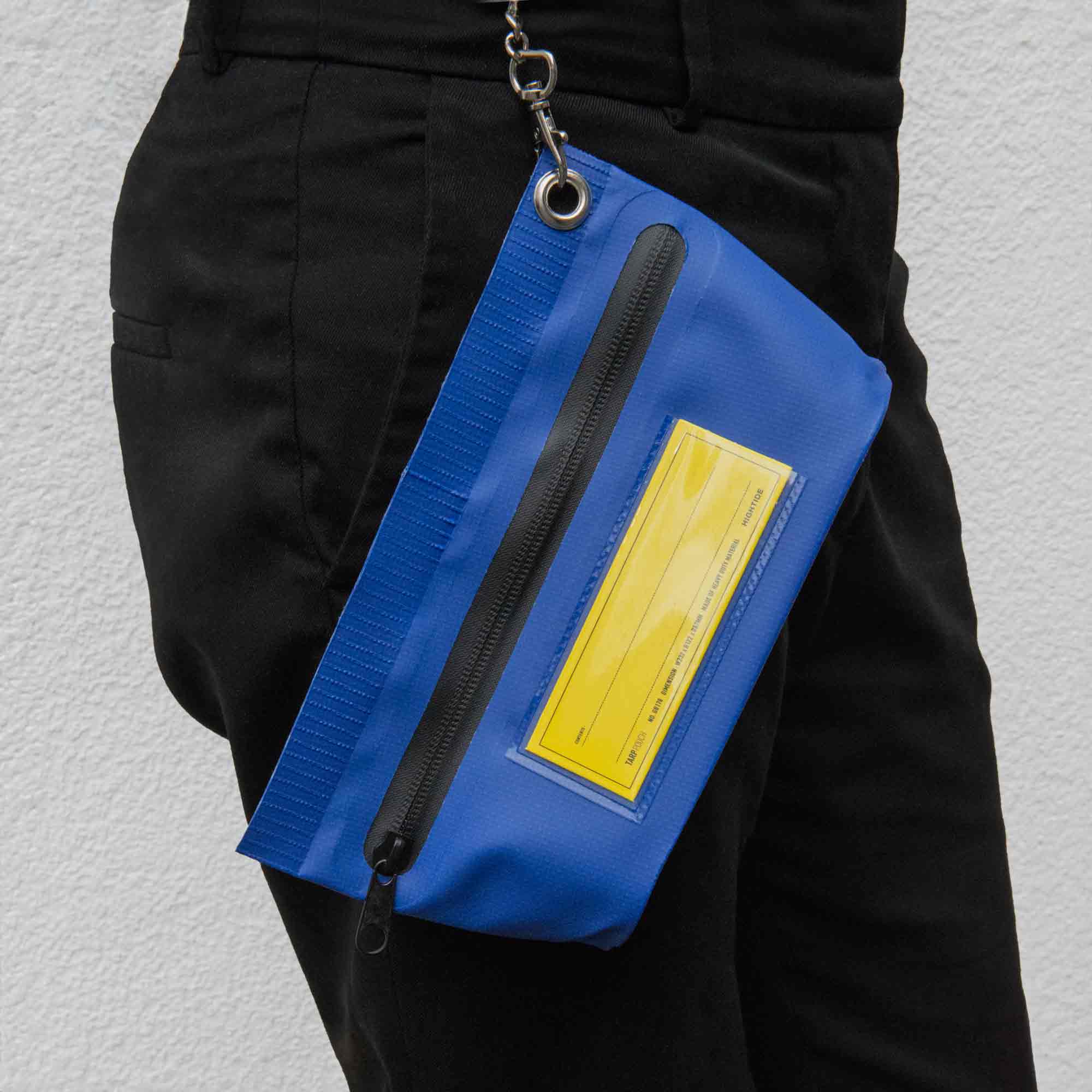 Eine Person in schwarzer Hose trägt eine Tarp Pouch S von Penco – blau mit gelbem Etikett und kleinen Karabinerhaken – aus strapazierfähigem Planenmaterial. Sie liegt an ihrer Seite, im Hintergrund eine strukturierte weiße Wand.