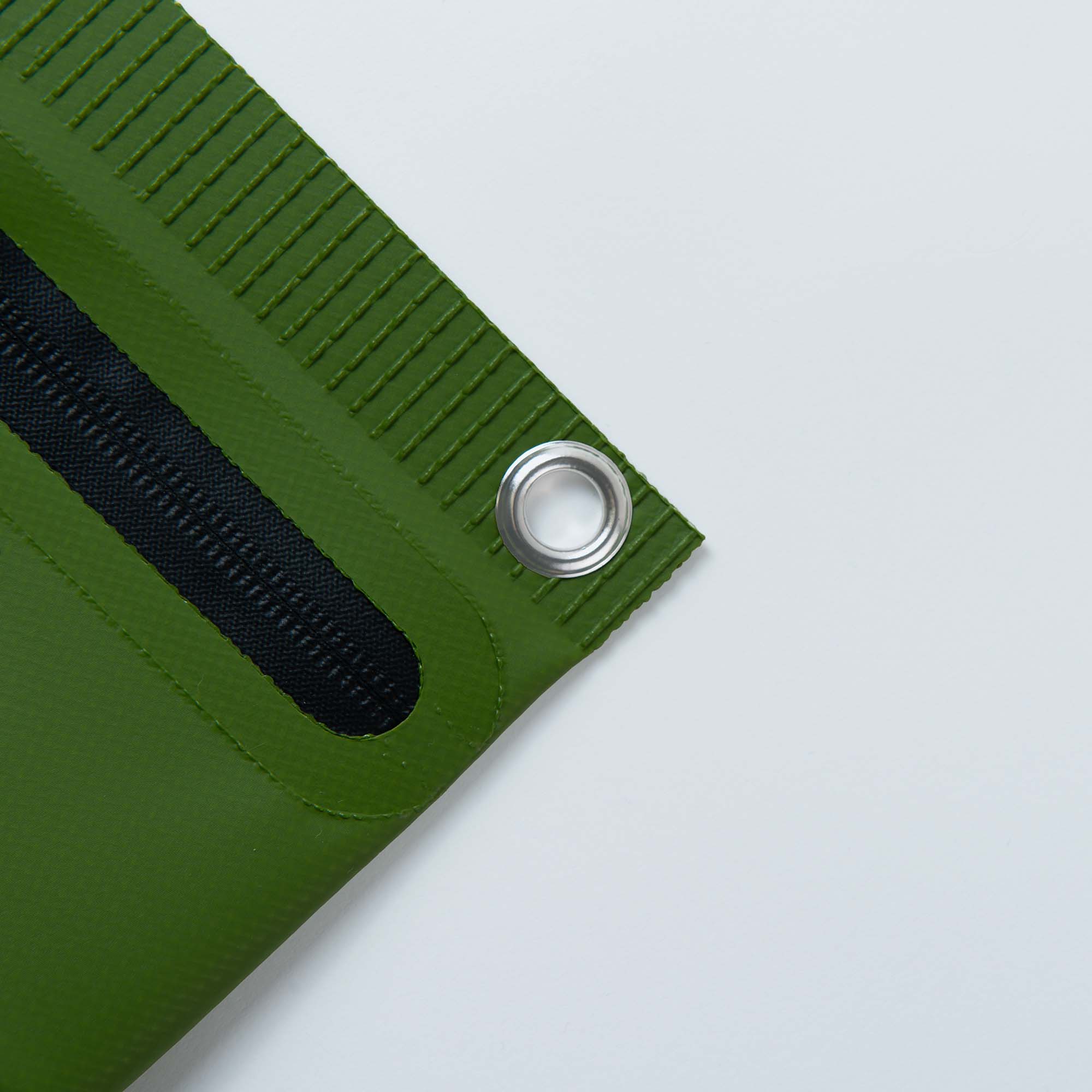 Nahaufnahme der Tarp Pouch S Corner von Penco aus strapazierfähigem Planenmaterial, mit schwarzem Streifen und glänzender Metallöse. Der strukturierte grüne Stoff kontrastiert mit dem glatten, hellen Hintergrund.