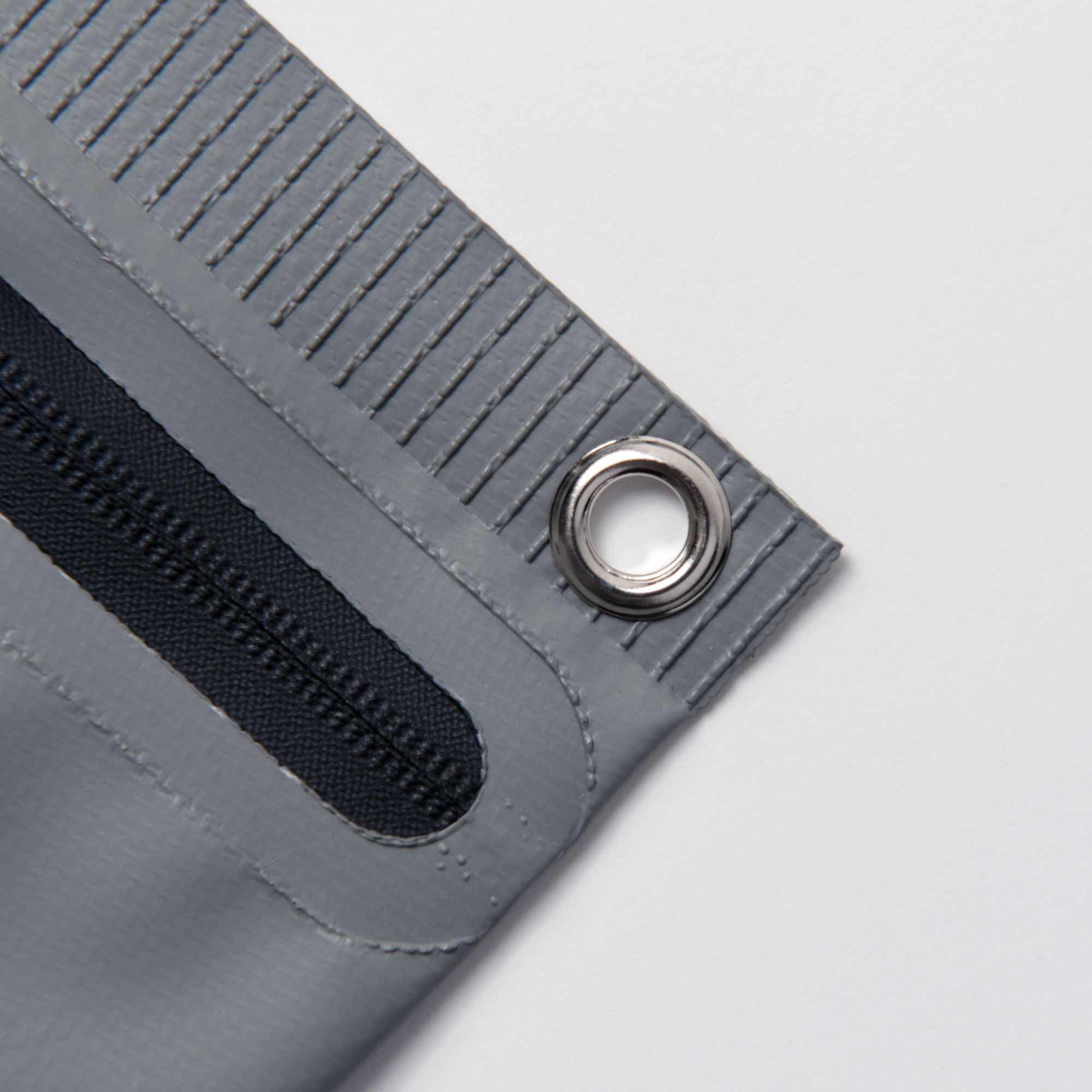 Eine Nahaufnahme der Tarp Pouch S von Penco zeigt eine graue Ecke mit einer Metalltülle und einem schwarzen Riemen, wodurch das strapazierfähige Planenmaterial hervorgehoben wird. Sein strukturiertes Aussehen wird durch parallele Nähte entlang der Kante verstärkt, die sich von einem schlichten weißen Hintergrund abheben.