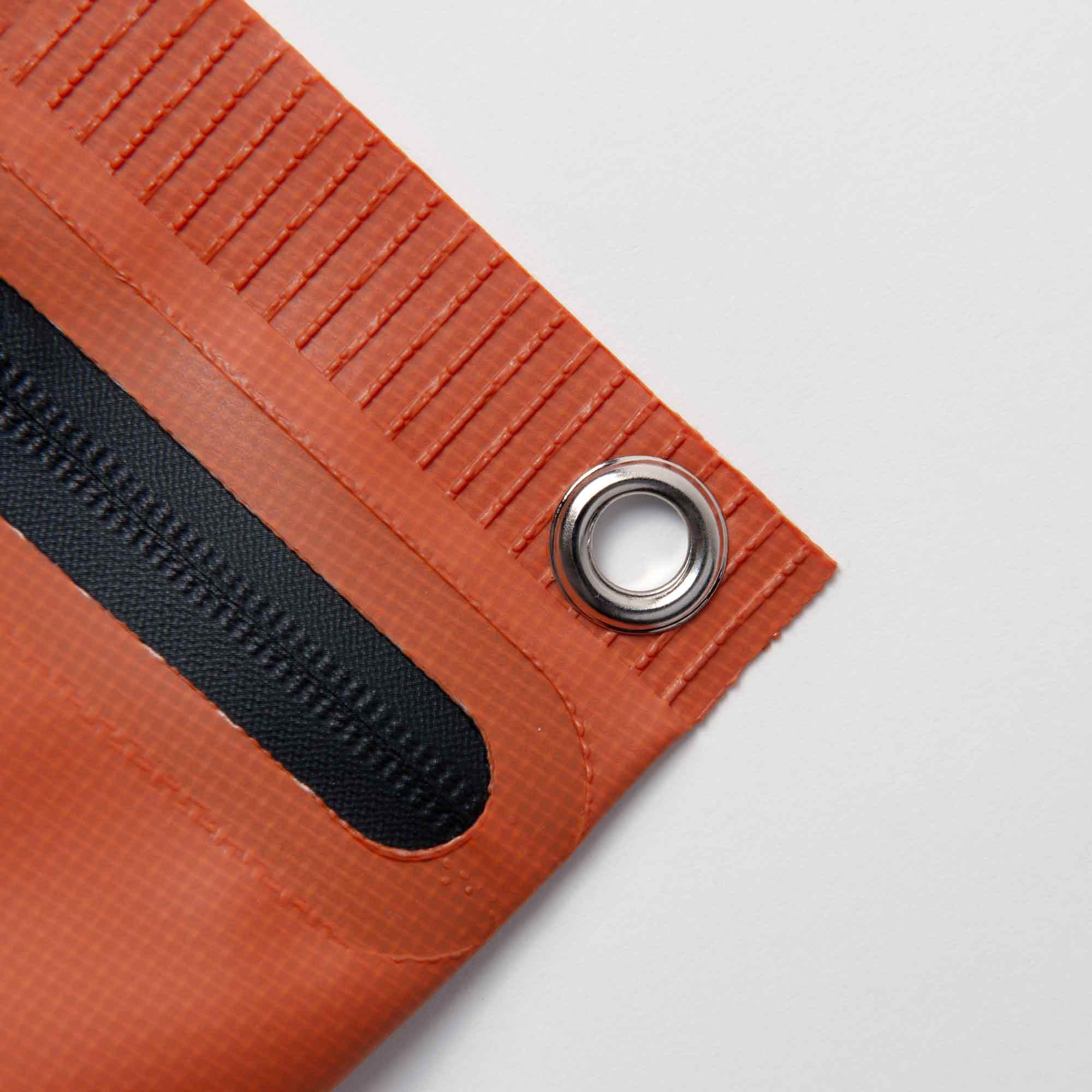 Nahaufnahme der Tarp Pouch S von Penco mit orangefarbener Ecke mit Metallöse und Karabinerhaken. Das Material wird durch einen schwarzen Streifen und sichtbare Randnähte auf weißem Hintergrund akzentuiert.