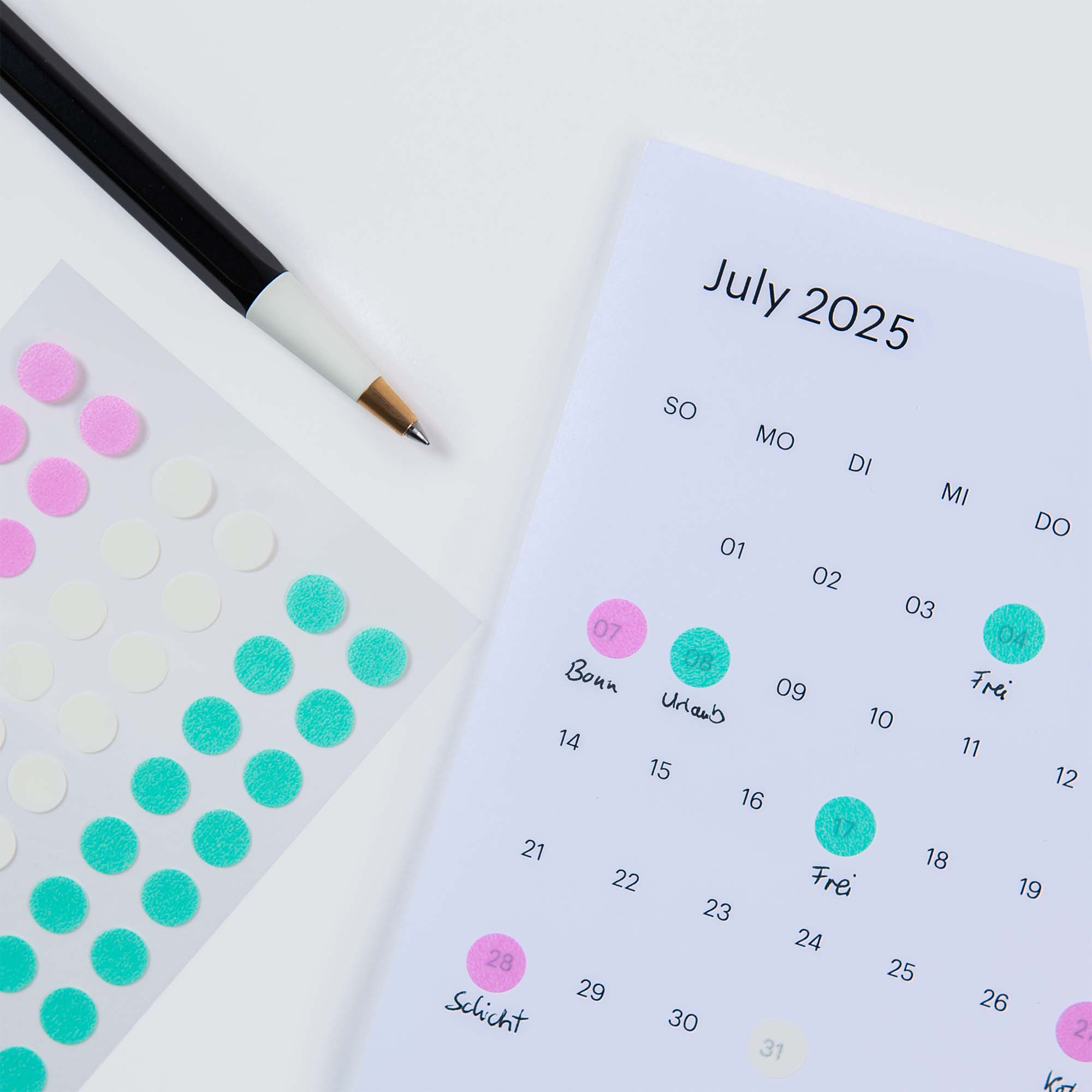 Ein Kalender für Juli 2025 auf einer weißen Fläche mit grünen und rosa Stalogy Circular Masking Tape Patches. In der Nähe liegen ein schwarzer Stift und ein Blatt mit Aufklebern aus hochwertigem japanischem Papier. Einige Daten zeigen handschriftliche Notizen.