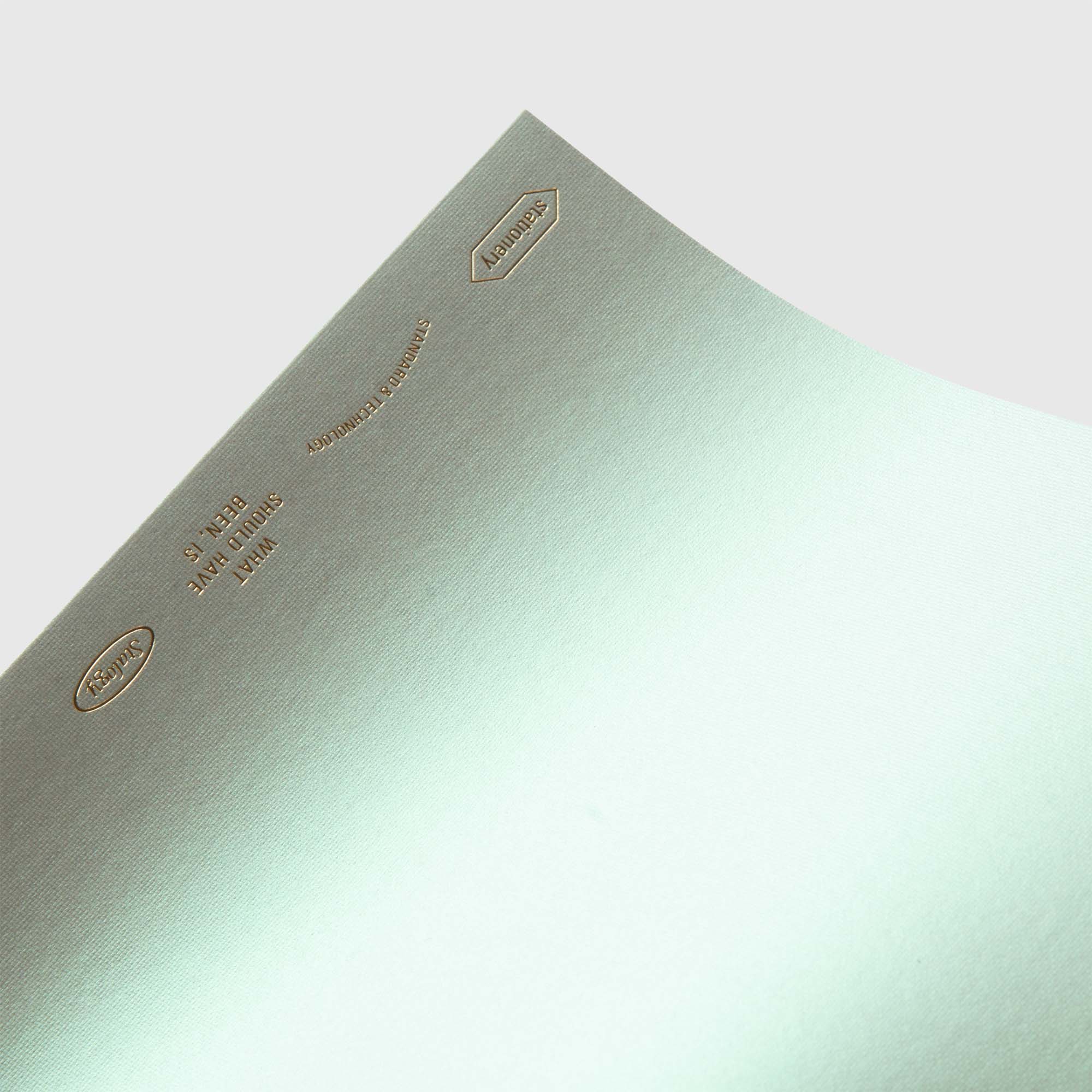 Eine Nahaufnahme von grünem Metallic-Strukturpapier mit goldgeprägtem Text und Logos, die einen polarisierenden Perlglanz-Effekt zeigen, aus der Stalogy Editor's Series 1/2 Year Notebook A5 Limited Edition, auf hellgrauem Hintergrund.