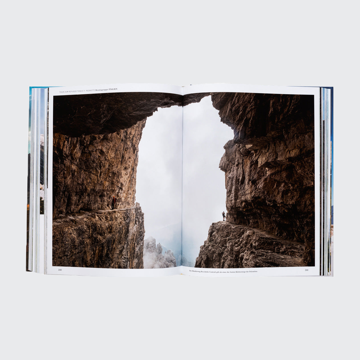 In einem offenen Buch aus dem Buch Wanderlust Alpen des Gestalten Verlags steht eine Person auf einer Klippe und fotografiert eine felsige, enge Schlucht mit steilen Klippen. Nebel oder Wolken füllen den Hintergrund, durch den der Himmel hindurchscheint und Alpenabenteuer und Fernweh verkörpert.