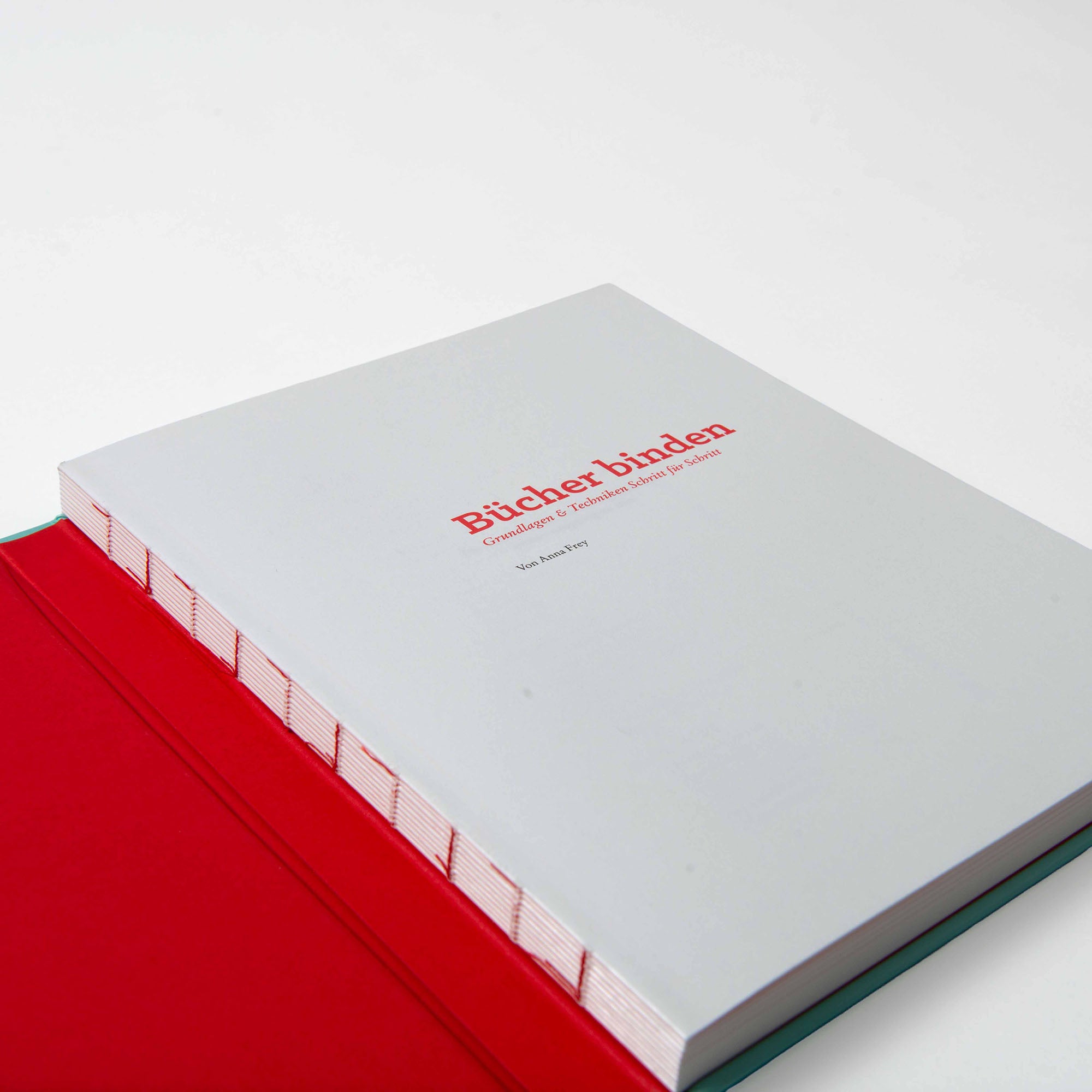 Anna Freys Buch "Bücher binden" mit schlichtem hellgrauem Einband und rotem Titel liegt aufgeschlagen auf rotem Grund und zeigt den sichtbaren, freiliegenden Buchrückeneinband und kleinere deutsche Untertitel.