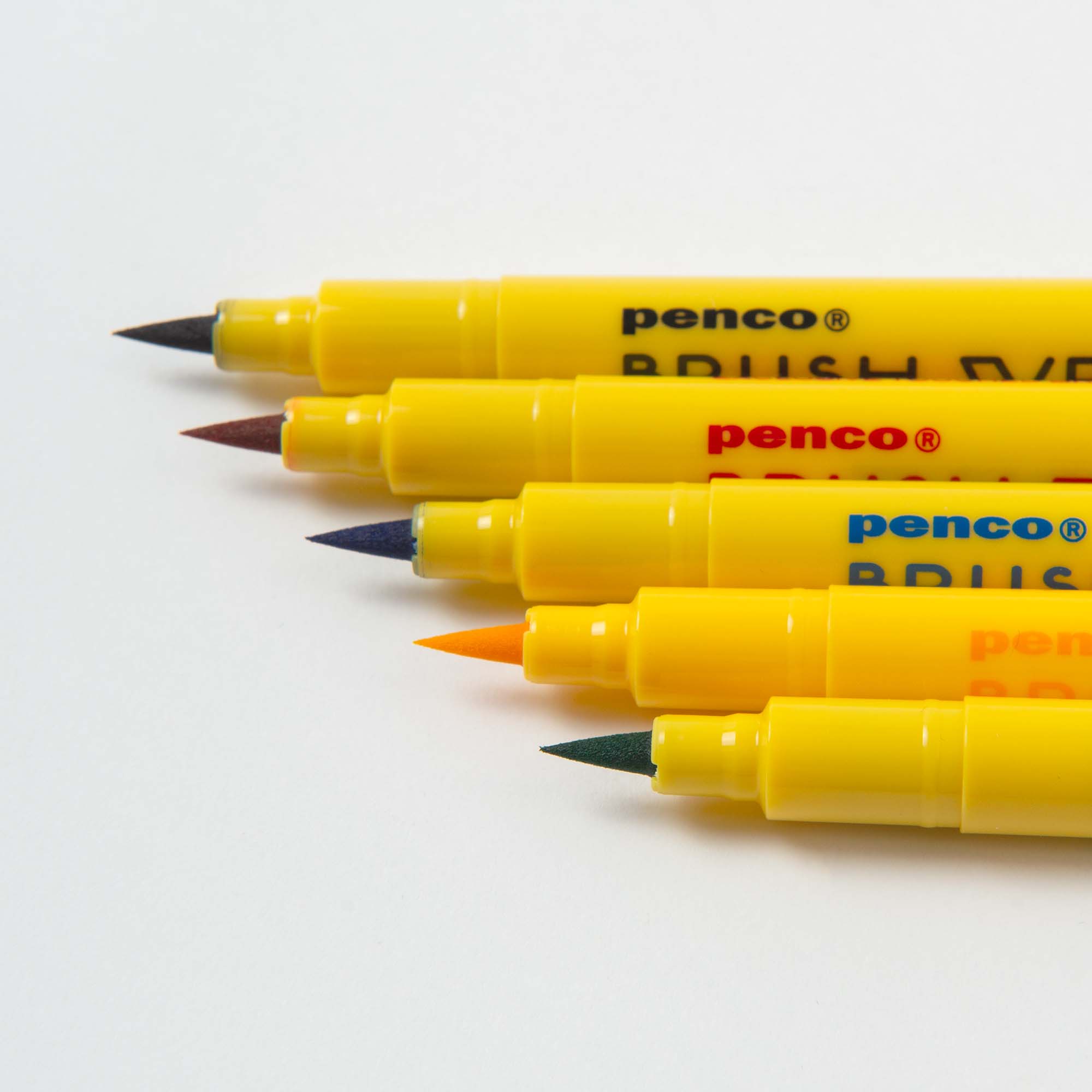 Fünf gelbe, doppelendige Penco Brushwriter-Set aus 5 Farben, Made in Japan, sind diagonal auf weißem Grund angeordnet, ihre farbigen Spitzen - braun, lila, blau, orange und grün - zeigen auf der linken Seite leuchtende Wasserfarben-Pigmente.