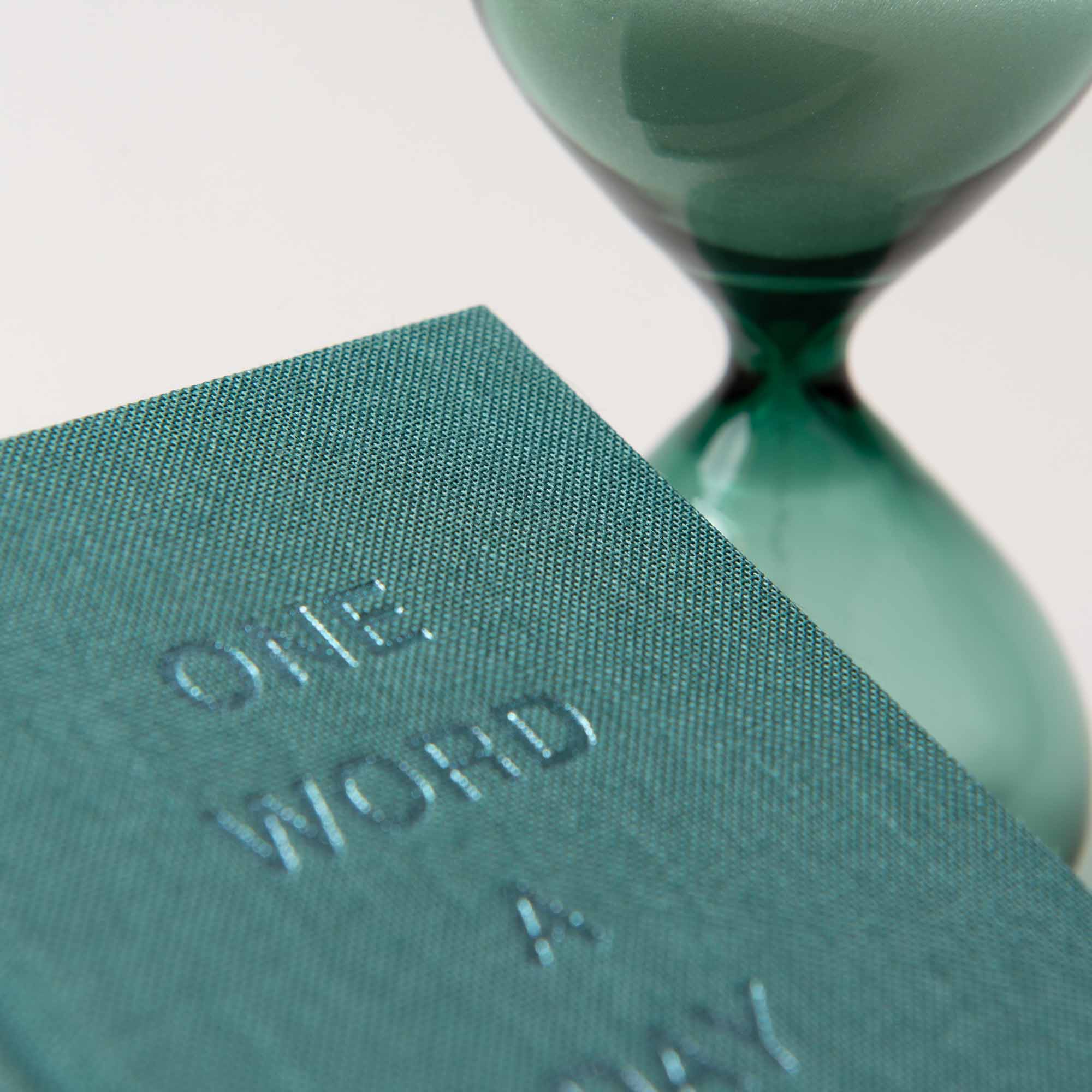 Nahaufnahme eines strukturierten grünen Buchumschlags mit der Prägung „One Word a Day mit Sanduhr“ von paperlux.store, neben einer handgefertigten Sanduhr. Die Komposition hebt die Textur des Buches und die eleganten Kurven der mit Sand gefüllten Sanduhr hervor und macht es zu einem idealen Geschenk.