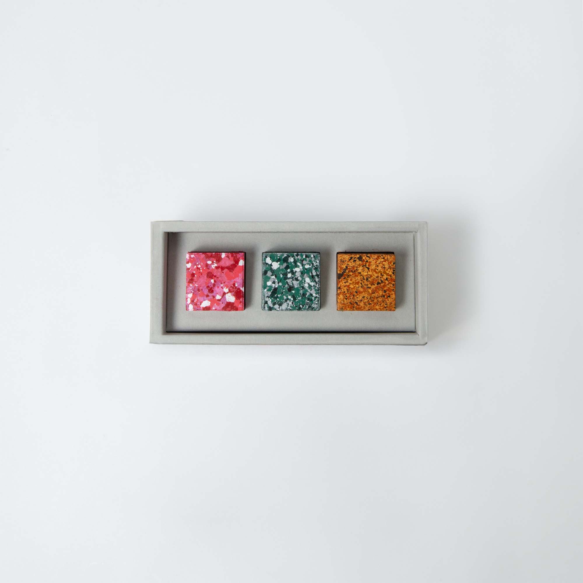 Das Set „Color Gem – das Wachsmalquadrat“ von Studio Unto besteht aus drei quadratischen Terrazzofliesen in ausgewählten Farbtönen – Rosa, Grün und Orange – mit einem gesprenkelten Muster, präsentiert in einer rechteckigen Box auf weißem Hintergrund.